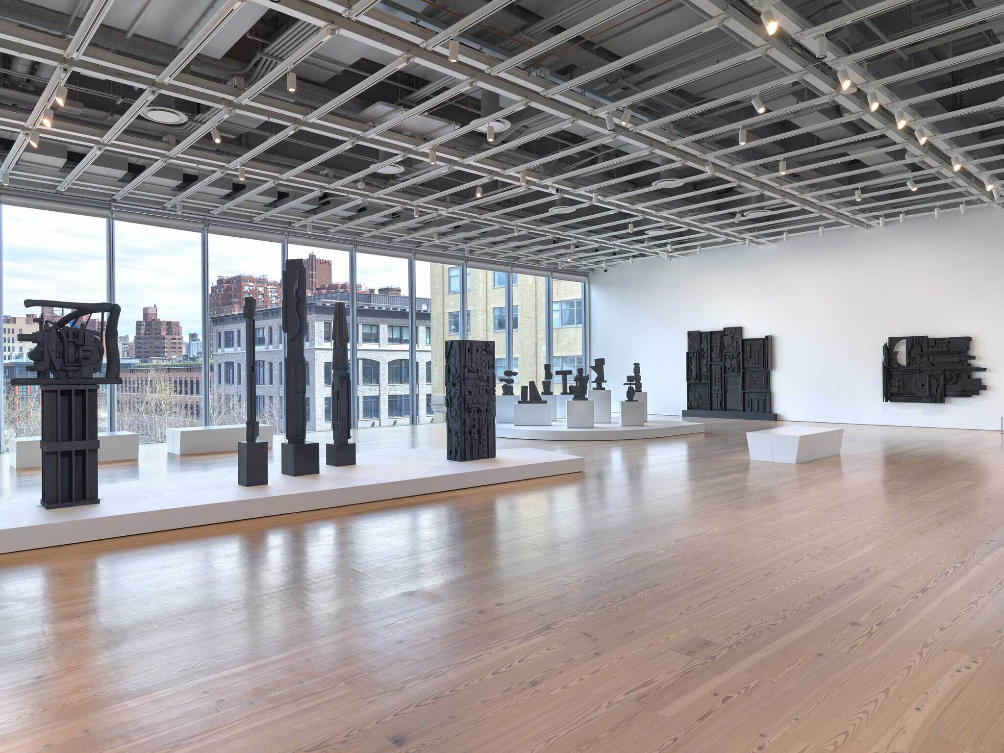 Modern art gallery with large windows, displaying several dark abstract sculptures on platforms and pedestals.