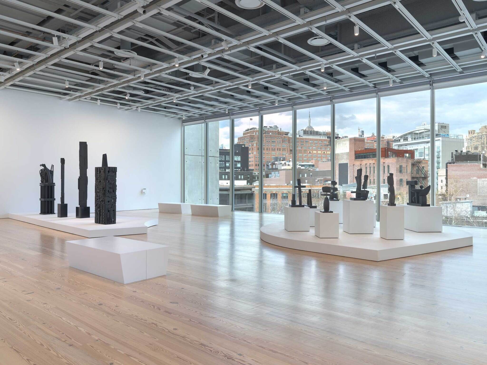 Modern art gallery with large windows, displaying several black abstract sculptures on white platforms and pedestals.