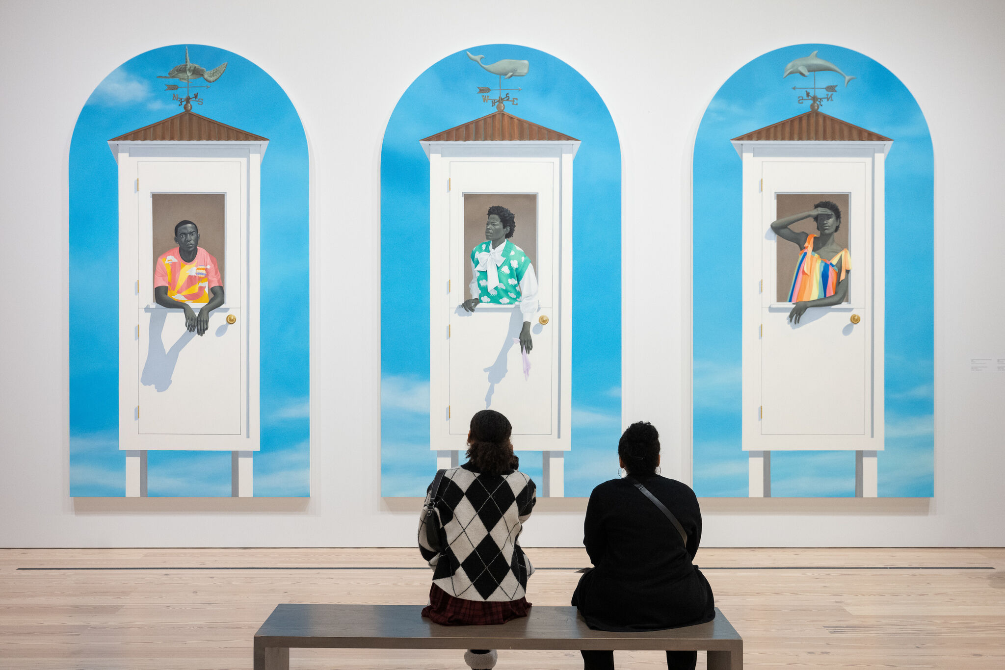 Two people sit on a bench looking at three large paintings of people leaning out of white doors.
