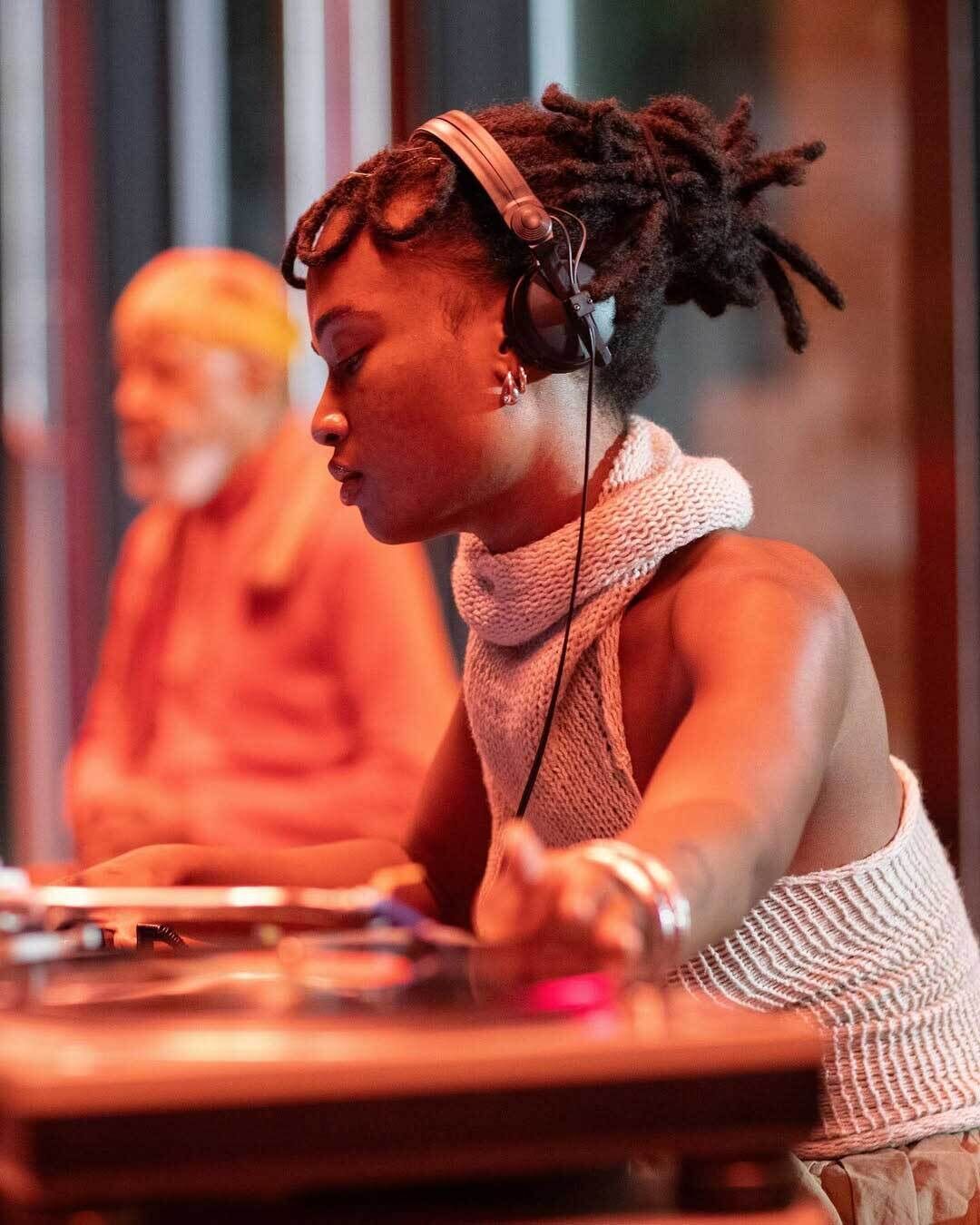 A woman wearing headphones is using DJ equipment, focused on adjusting controls, with another person in the background.