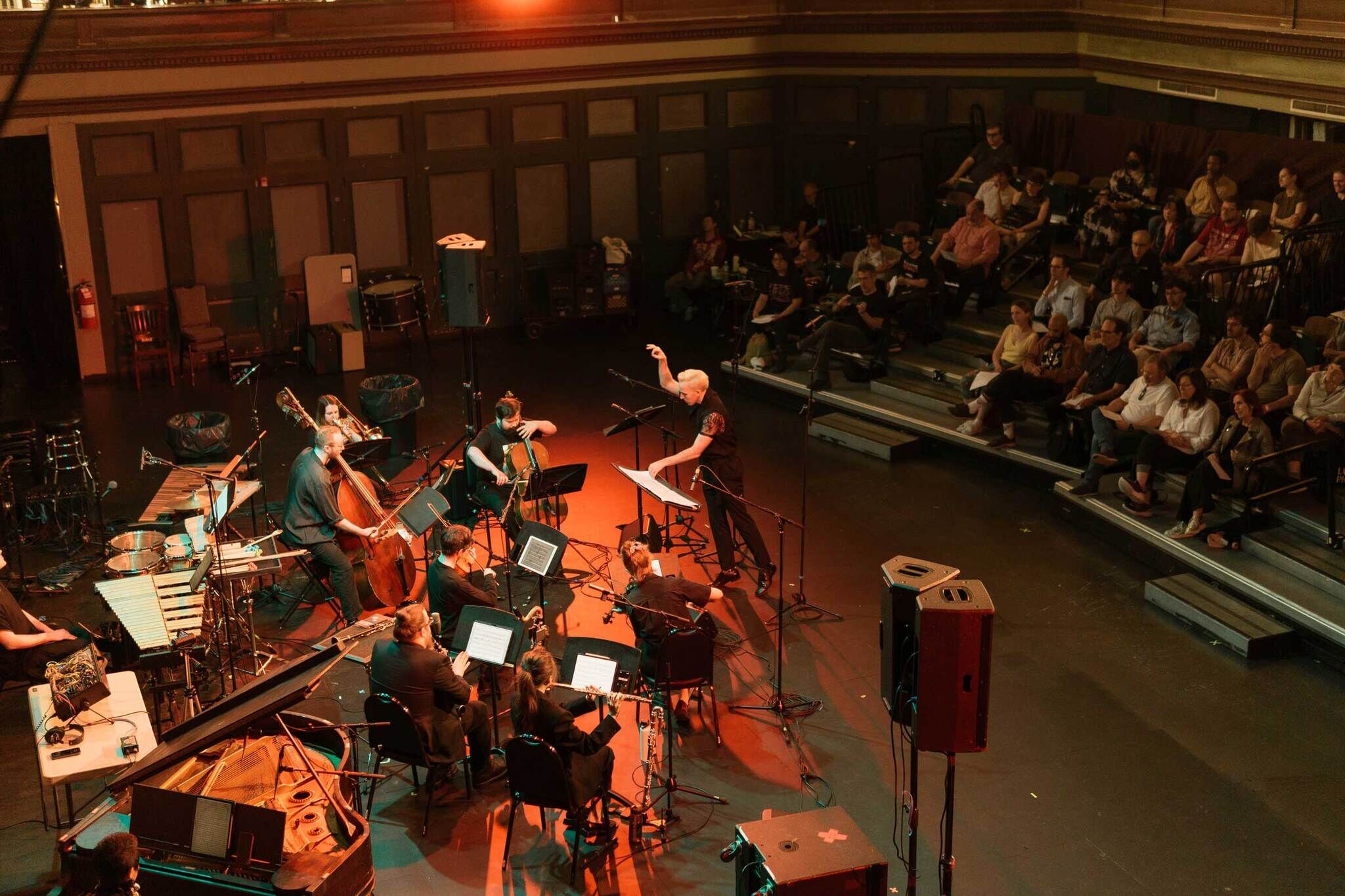 A conductor leads a small orchestra as people sit in bleachers watching the performance in a dimly lit hall.