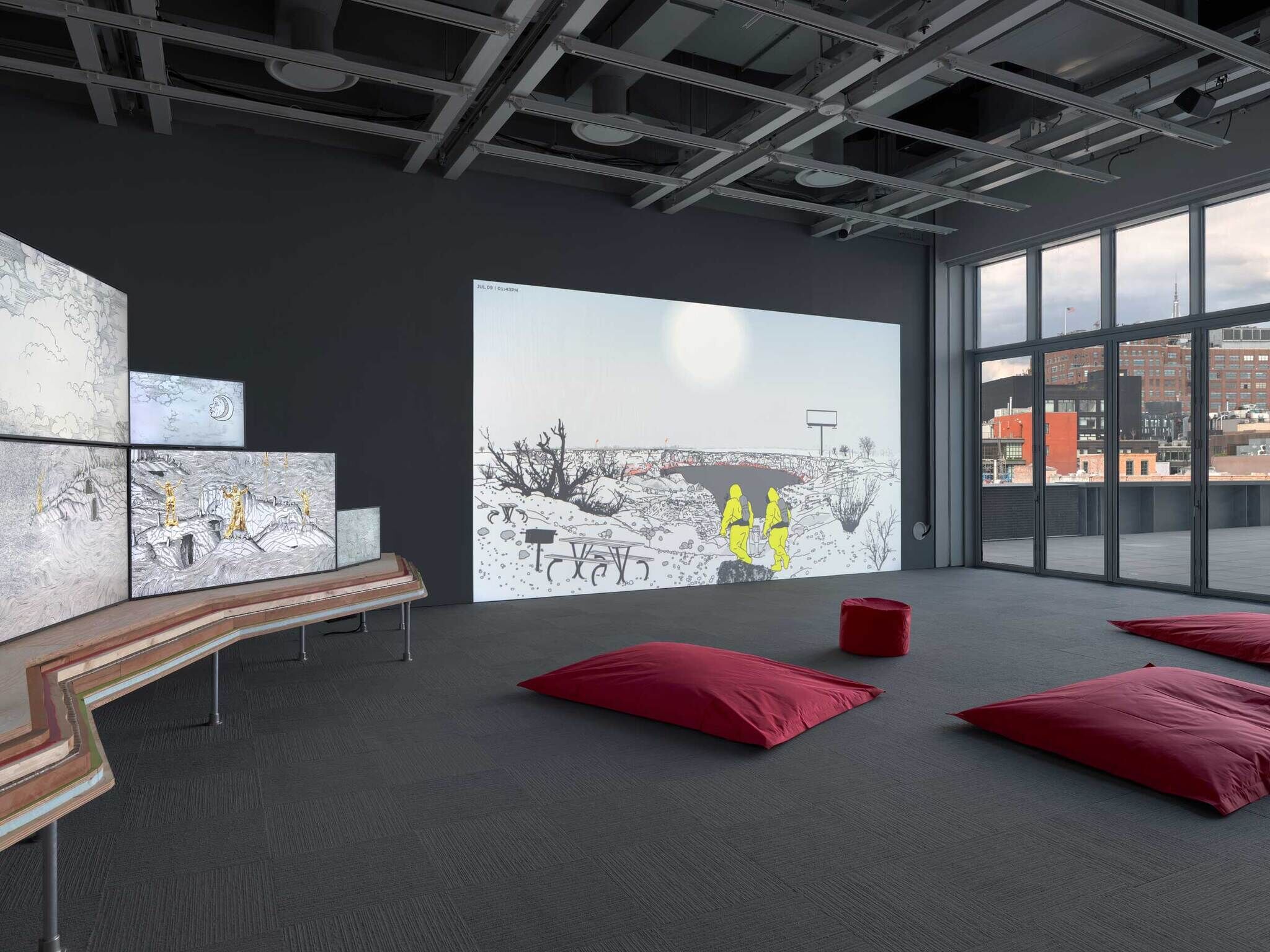 Large room with digital art on the walls, red floor cushions, and big windows showing city buildings outside.