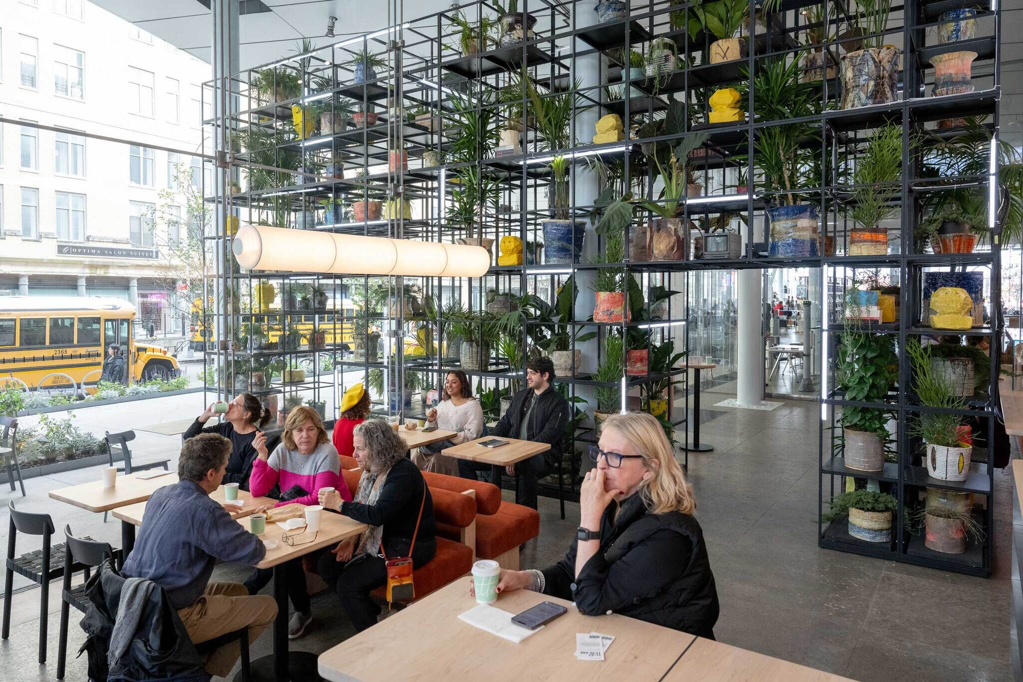 People sit at tables in a bright café with large shelves of potted plants and school buses outside the window.