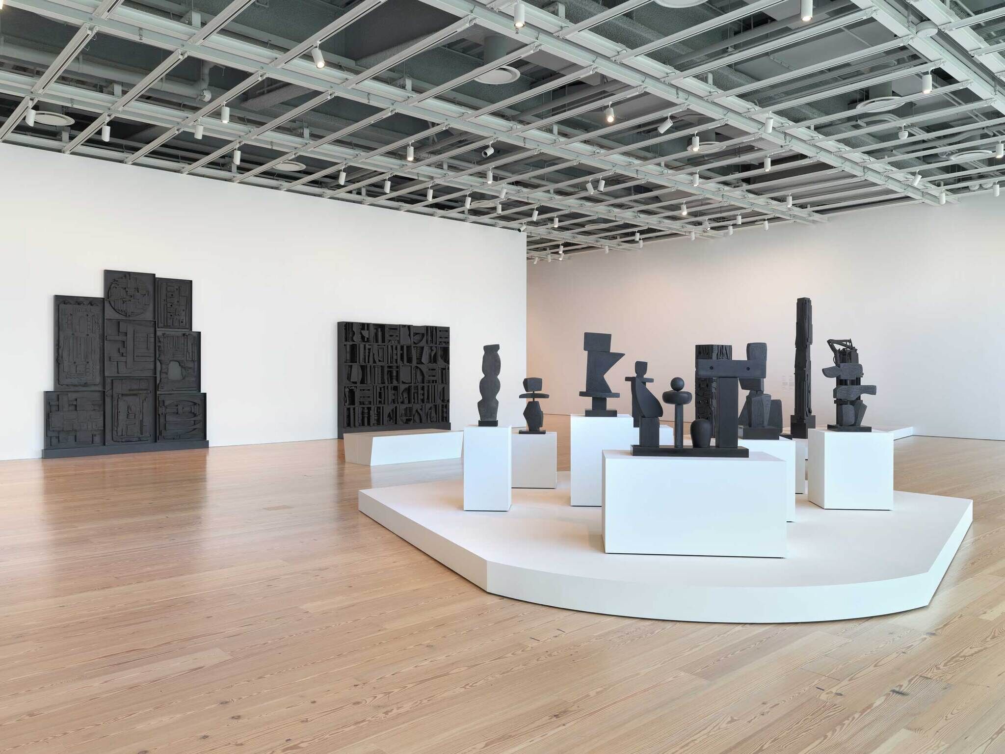 A modern art gallery full of black sculptures made of found materials. Two larger black sculpture are flush against the wall on the left and multiple smaller black sculptures of various shapes and sizes are put on white pedestals closer to the foreground.