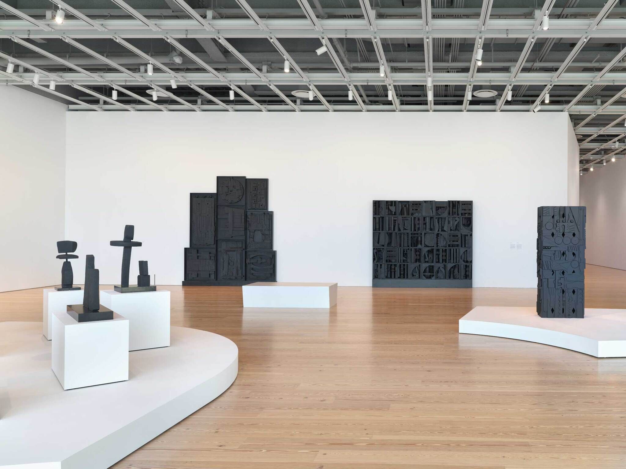 A room full of black sculptures that are made out of found materials. In the background, the two largest black sculptures are flush against the white wall. In the foreground, various smaller black sculptures are on white pedestals.