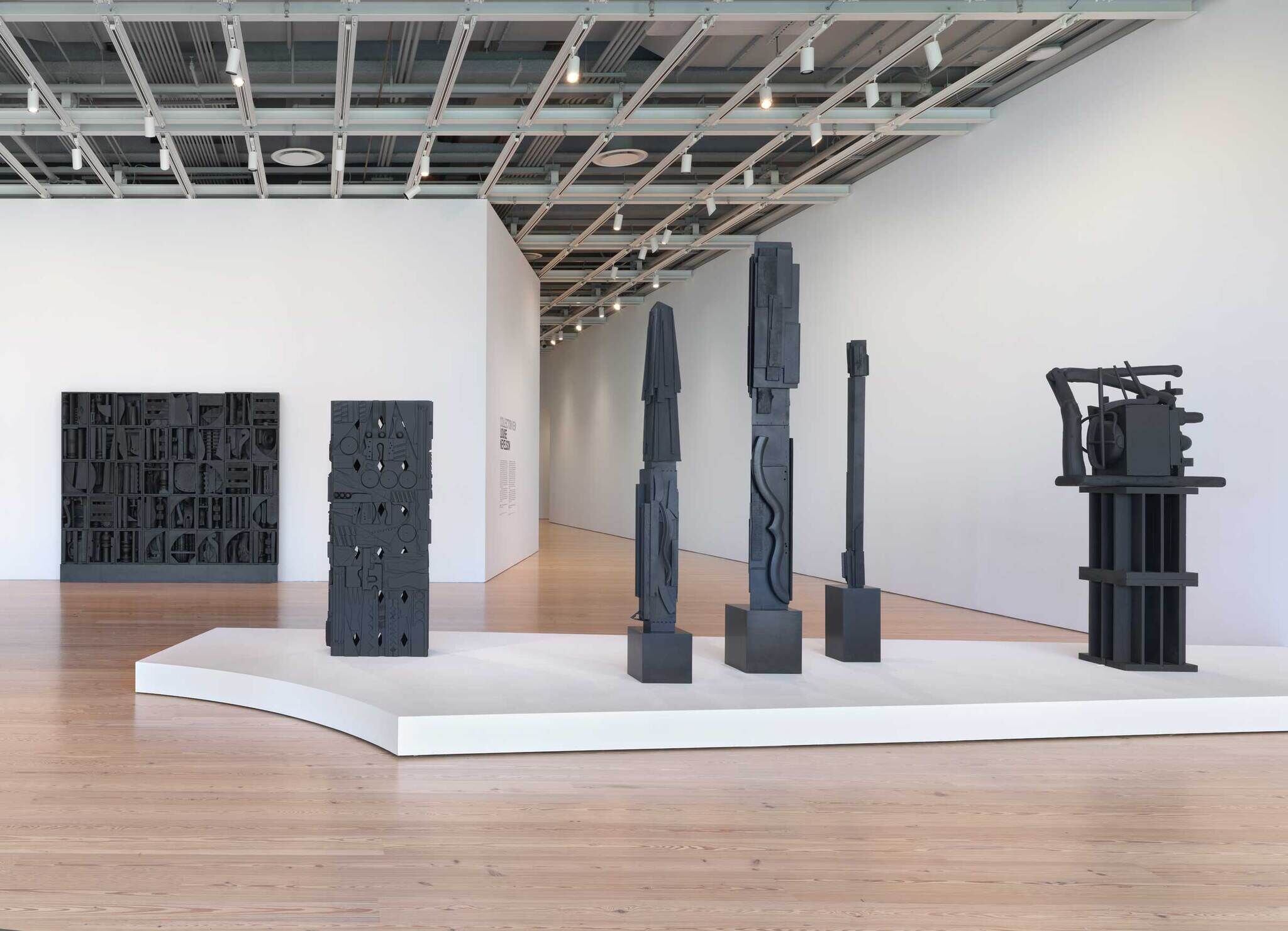 Several tall, dark abstract sculptures and textured panels are displayed on a white platform in a modern gallery.