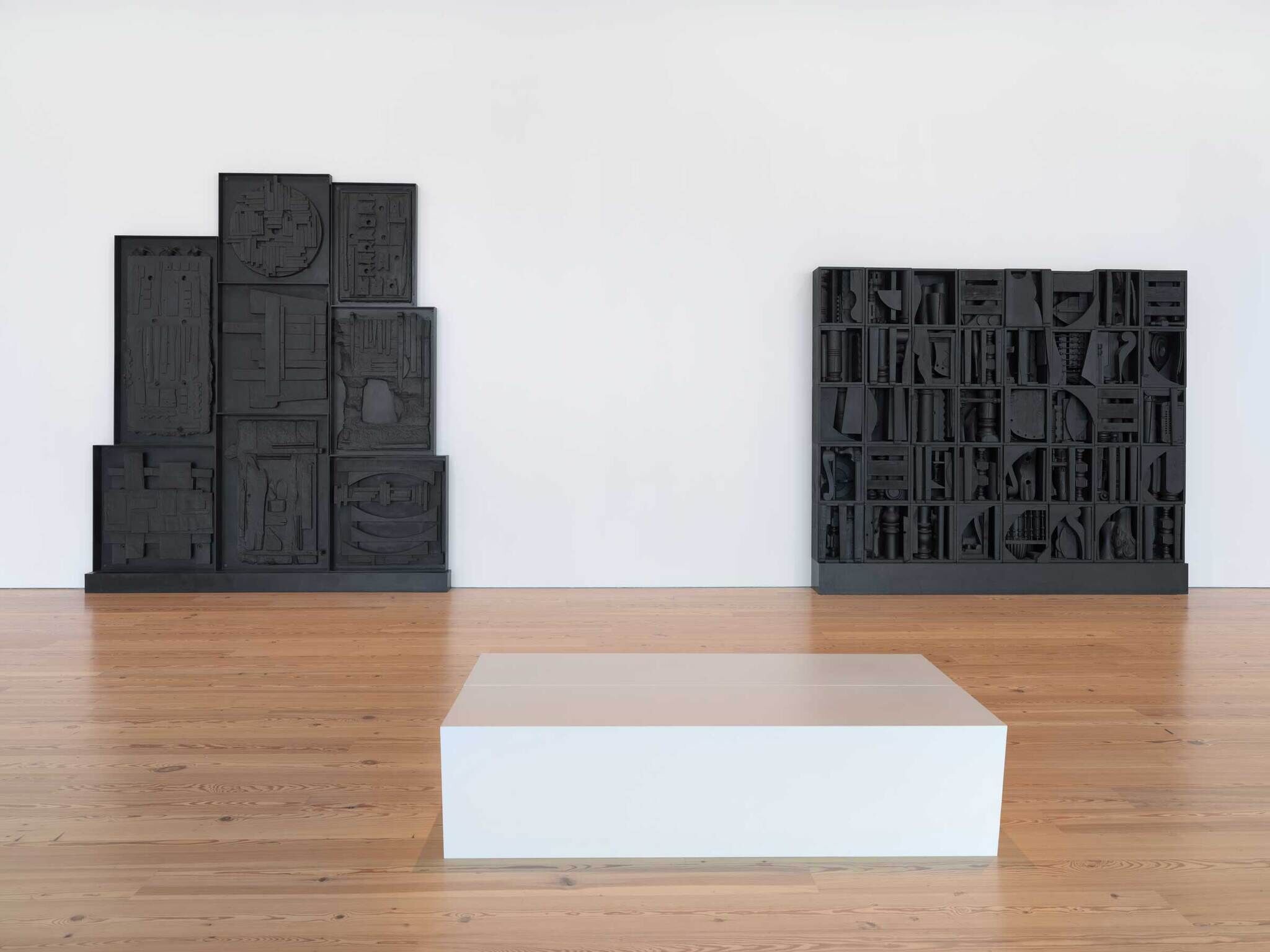 Two big black sculptures made out of found objects against a white wall. In the foreground there is a rectangular white bench.