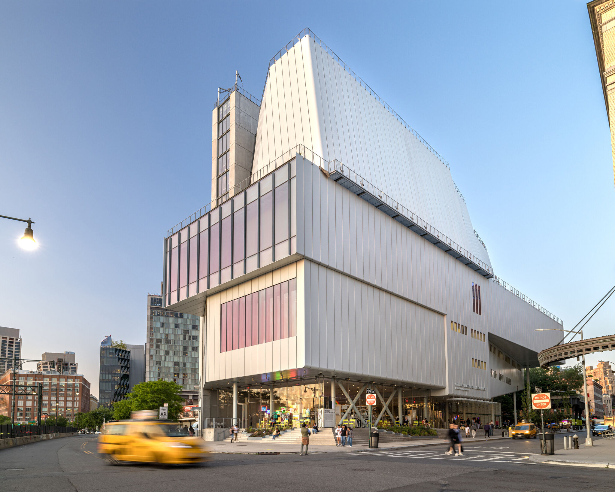 Large modern building with angled white panels, people walking outside, and yellow taxis driving by on the street.