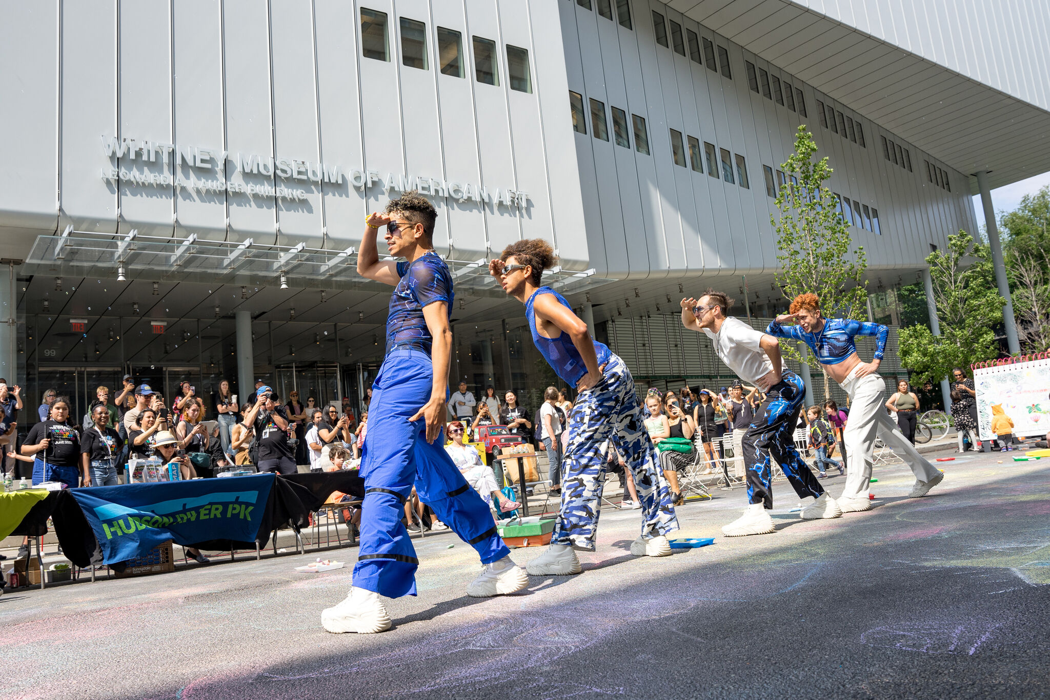 Four dancers perform in colorful outfits outside the Whitney Museum of American Art, with a crowd watching them.