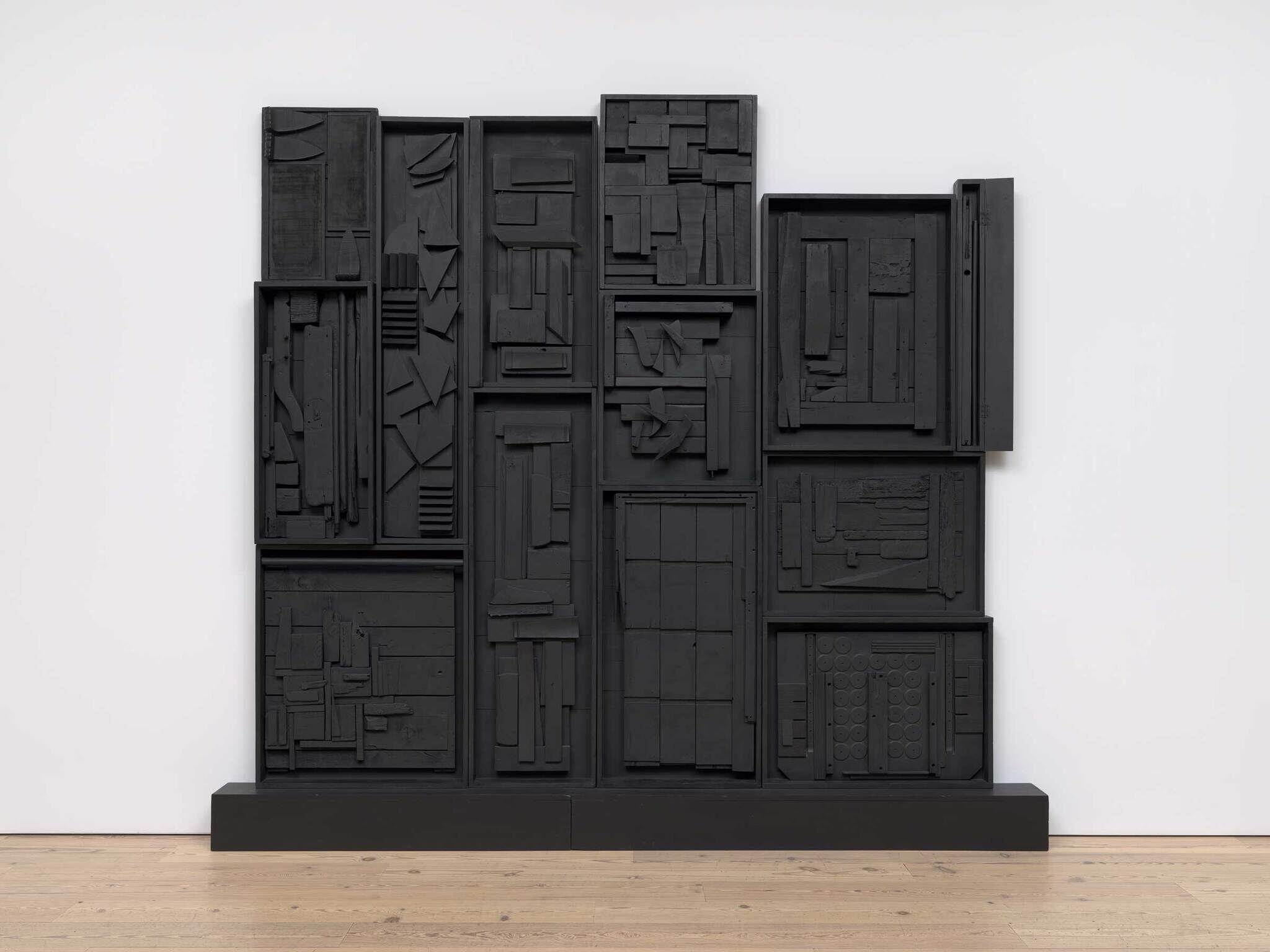 Large black wall sculpture made of wooden pieces arranged in geometric patterns, divided into several rectangular sections.
