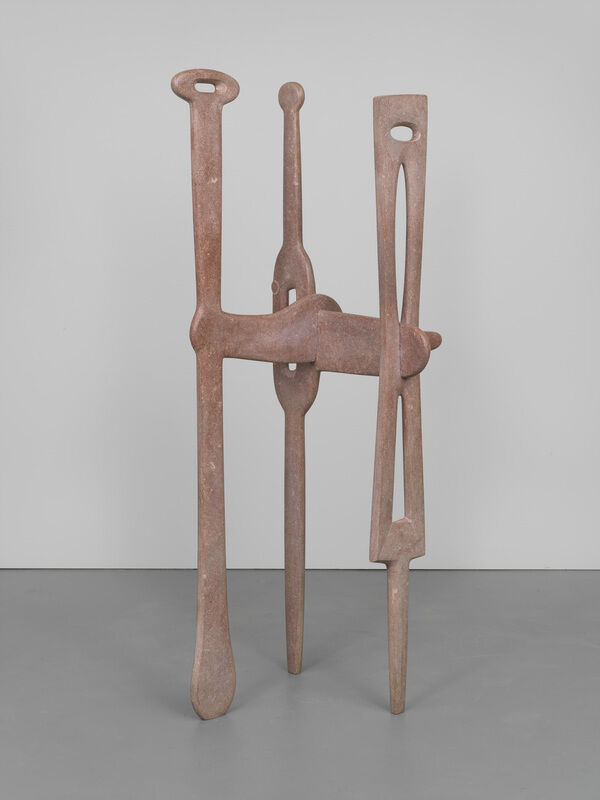 Tall abstract metal sculpture with three vertical, elongated shapes joined together, standing on a gray floor.