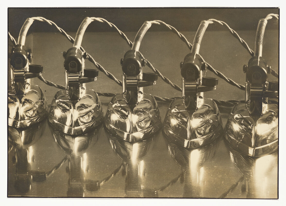 Five shiny electric irons are lined up in a row, with their cords twisted behind them.
