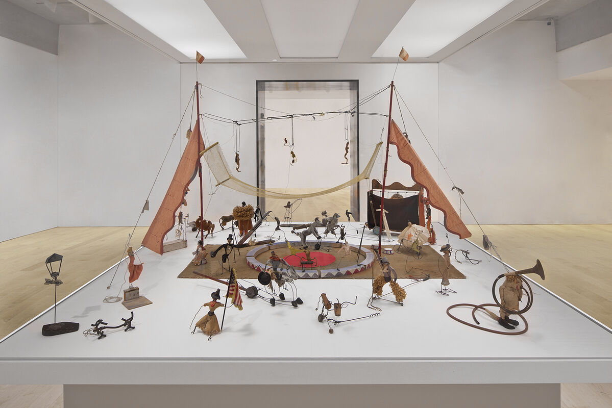 Miniature circus scene with wire and fabric figures performing acts under a tent, displayed on a white platform.