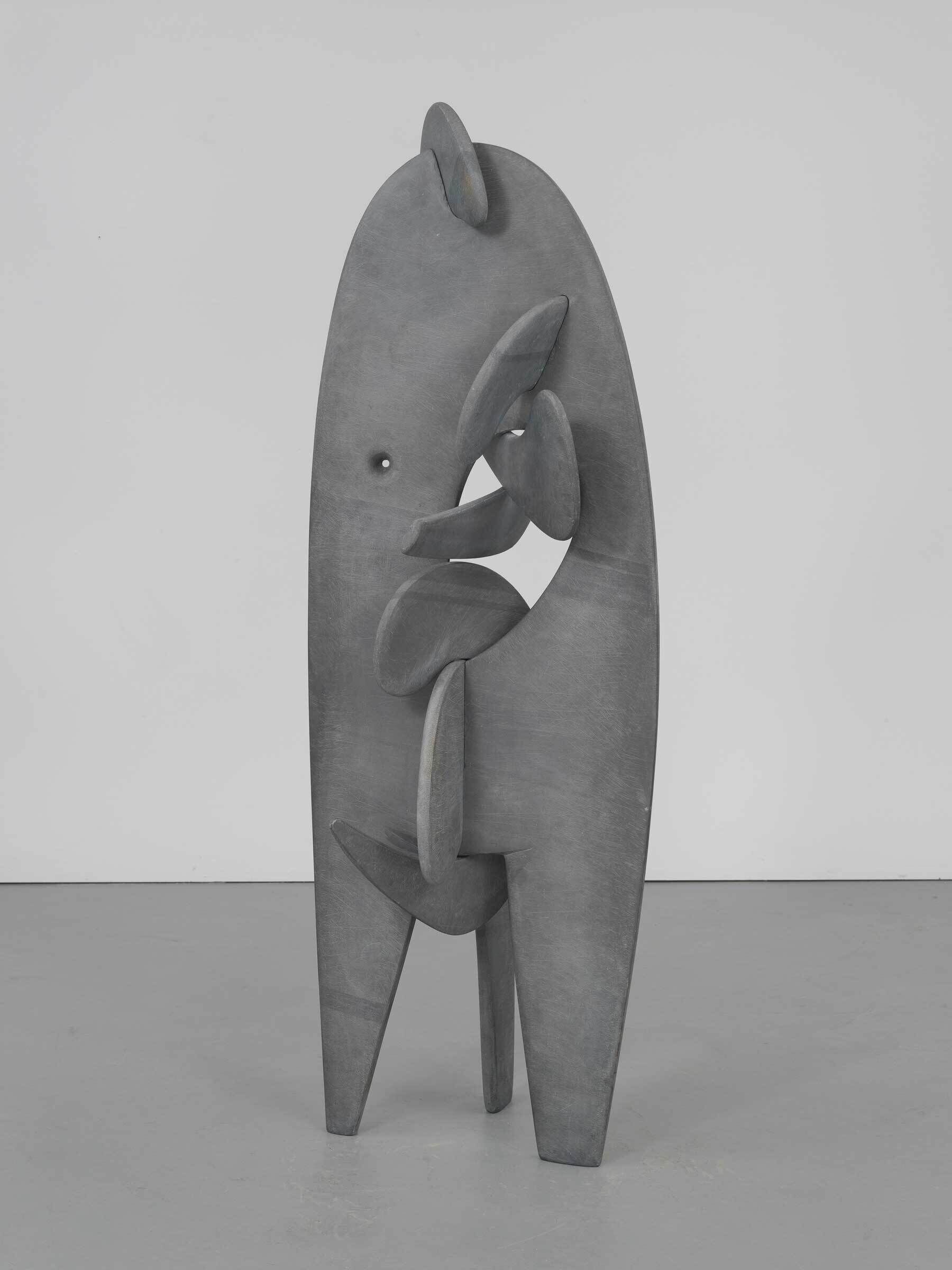Tall, abstract gray sculpture with rounded shapes and cutouts, standing on two legs in a plain room.