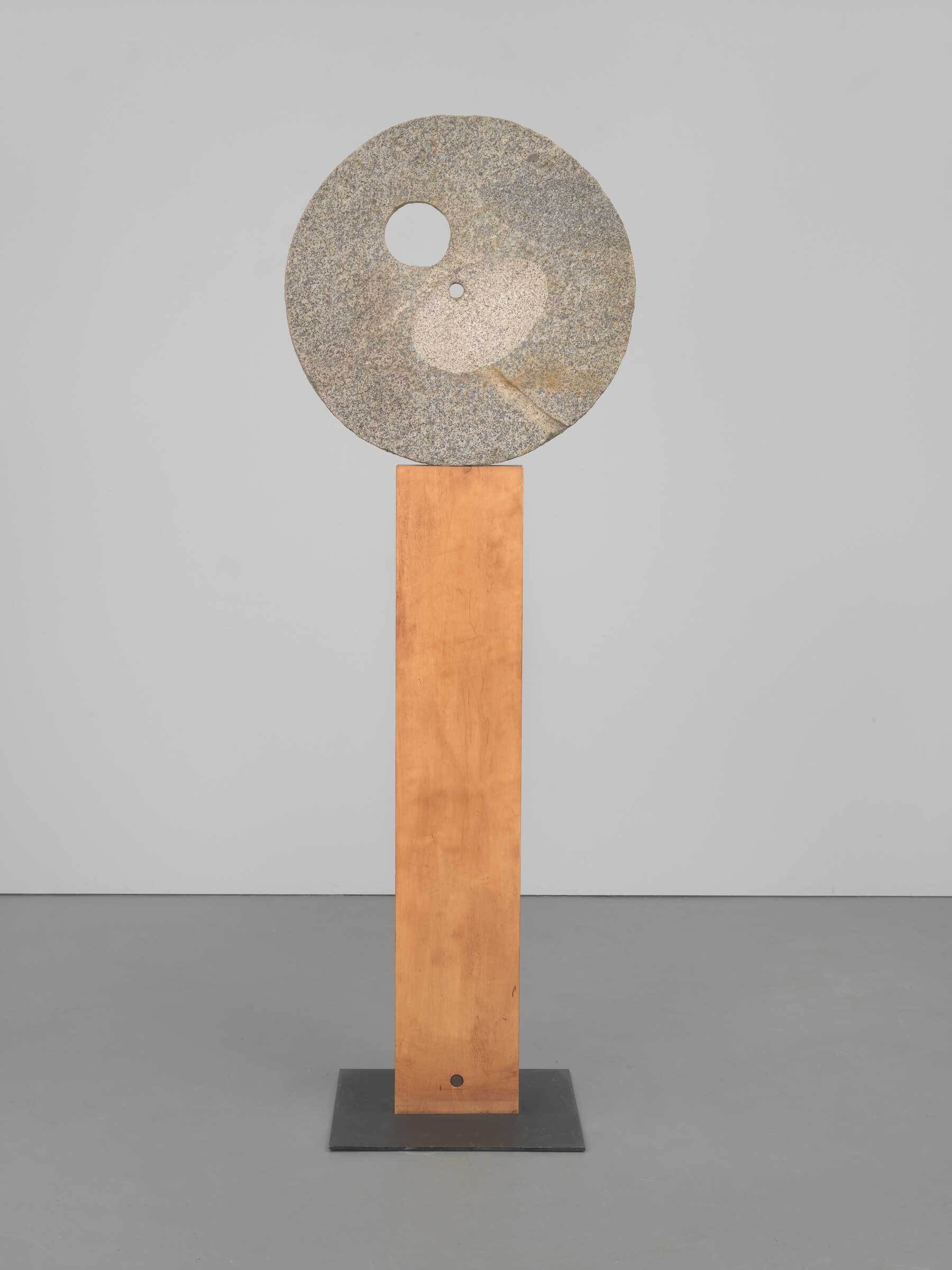 Tall sculpture with a round stone disk featuring two holes, mounted on a wooden rectangular base.