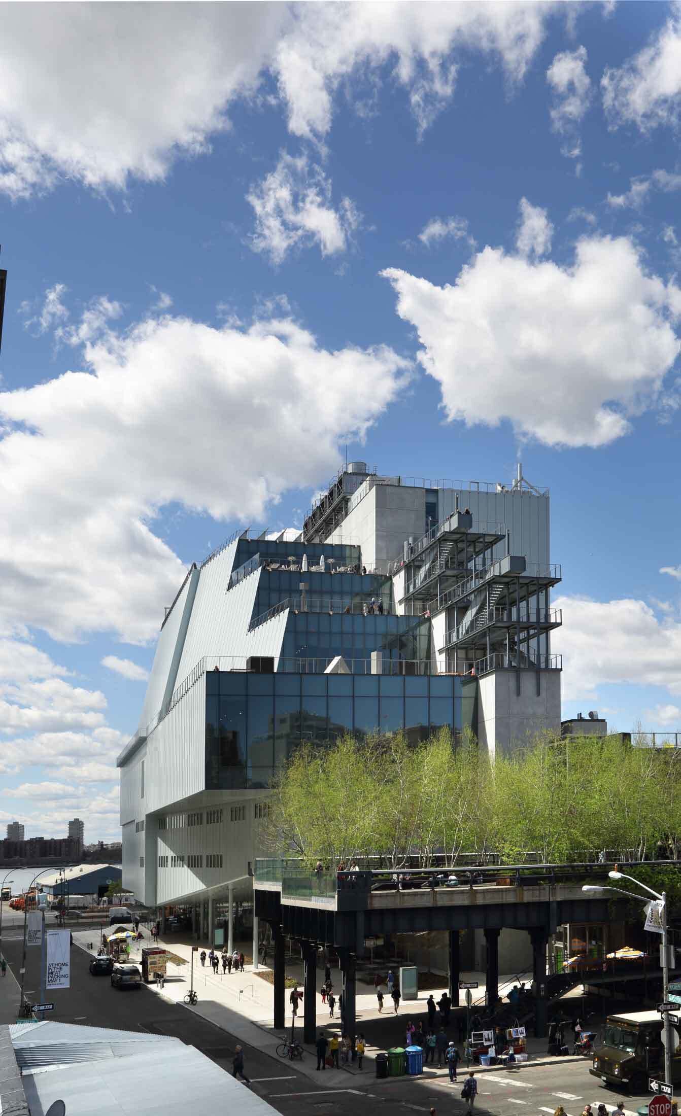 Modern glass and metal building with outdoor terraces, next to a raised park with people walking below and above.