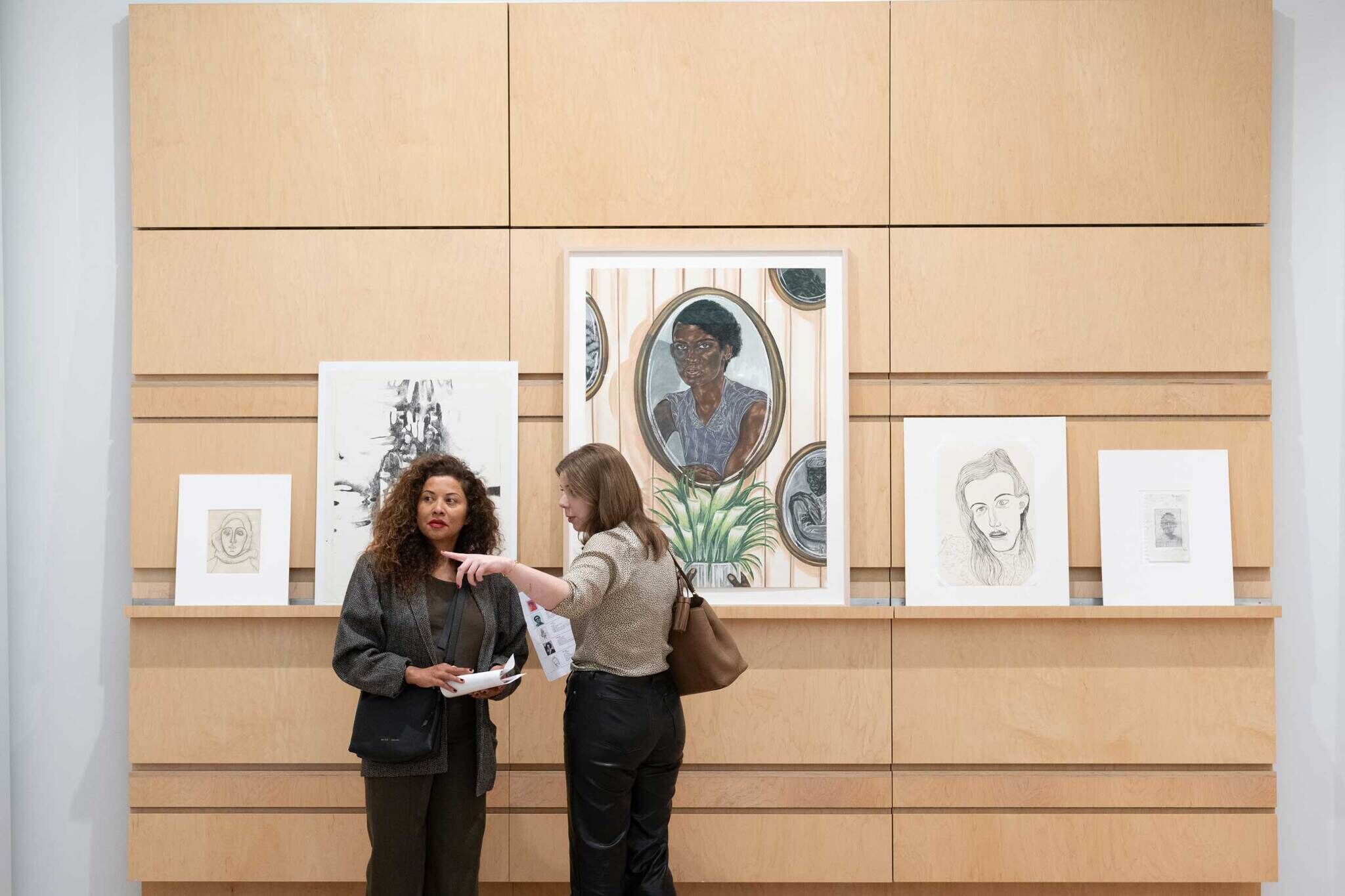Two women stand in front of portrait drawings and paintings; one woman points while talking to the other.