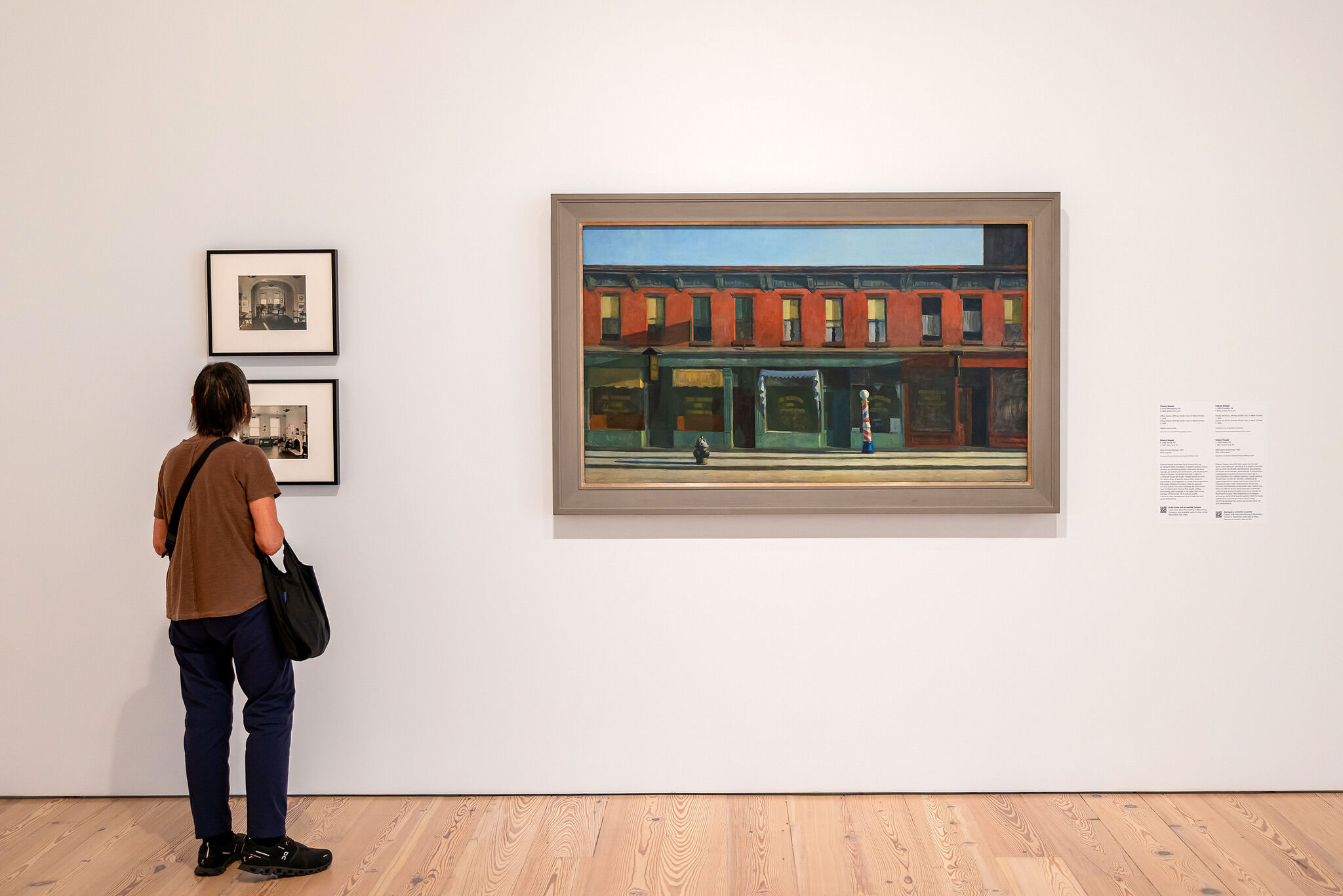 A person stands in a gallery looking at two small black-and-white photos next to a large painting of buildings.