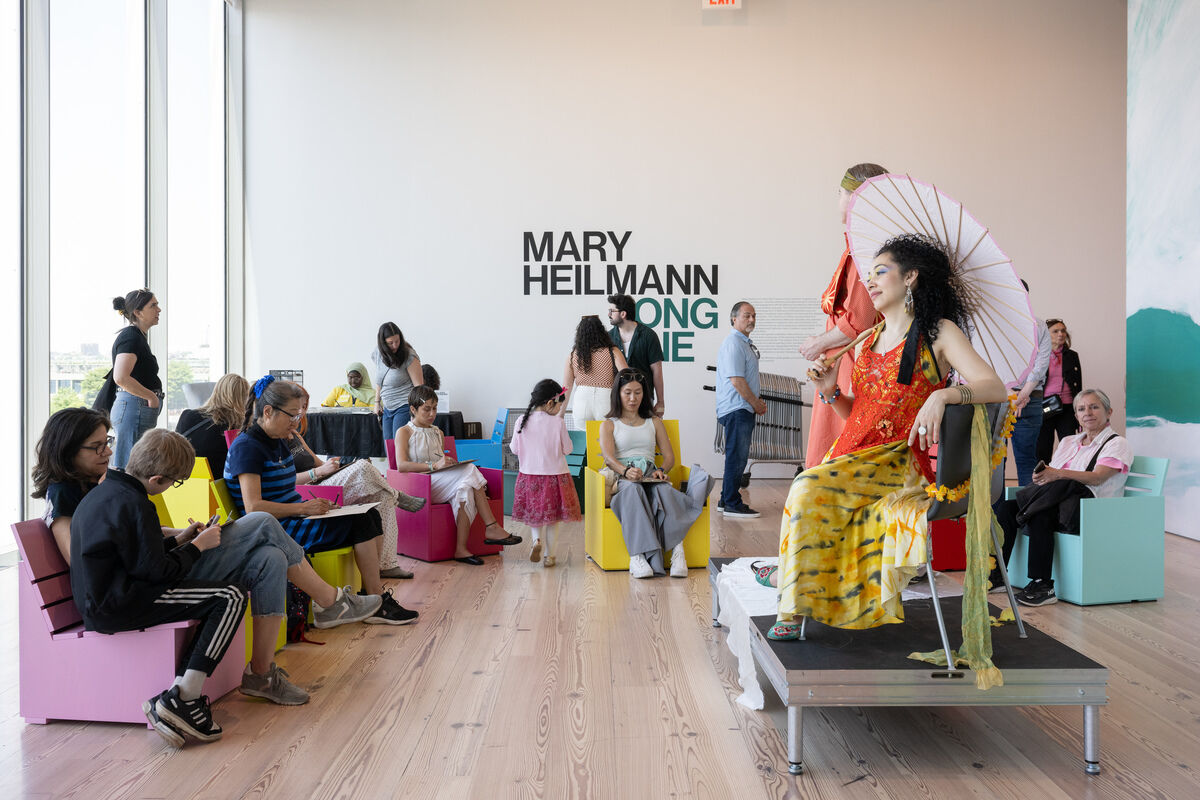 People sit on colorful chairs sketching a woman in a bright dress holding a parasol; “Mary Heilmann” text on wall.