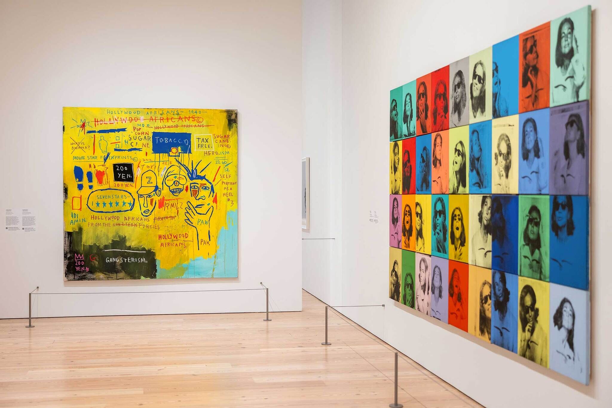 Two large modern art pieces hang in a gallery; one is colorful with text, the other has repeated portraits.
