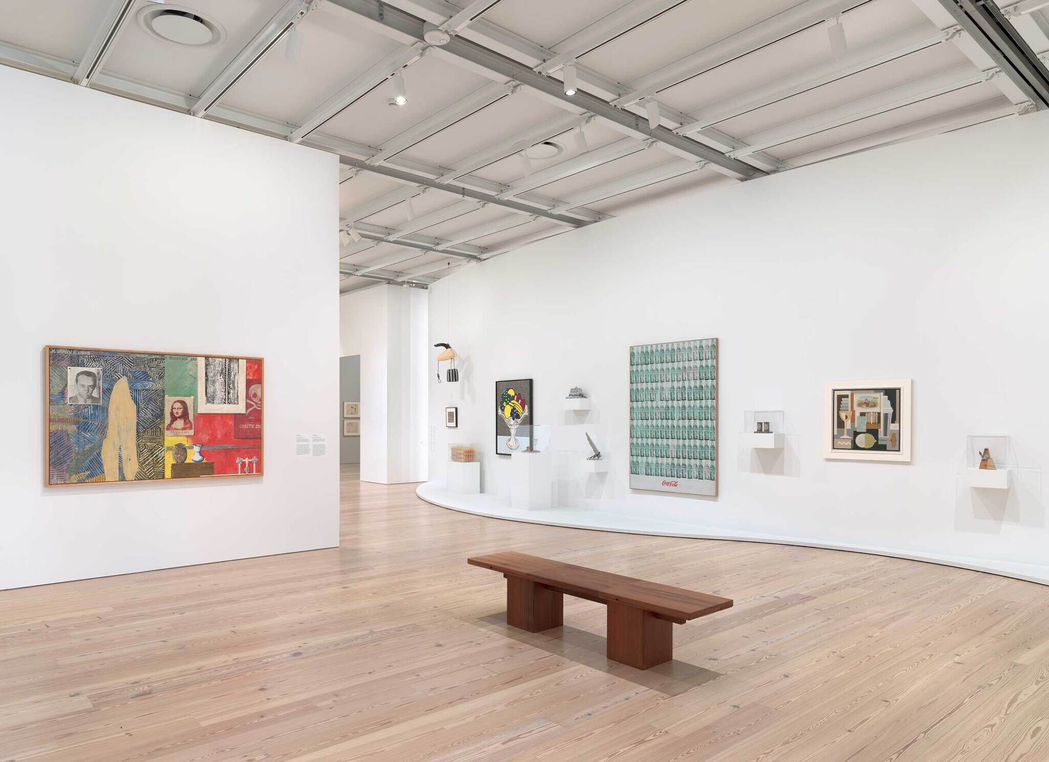 Art gallery with modern paintings and sculptures on white walls, and a wooden bench in the center of the room.
