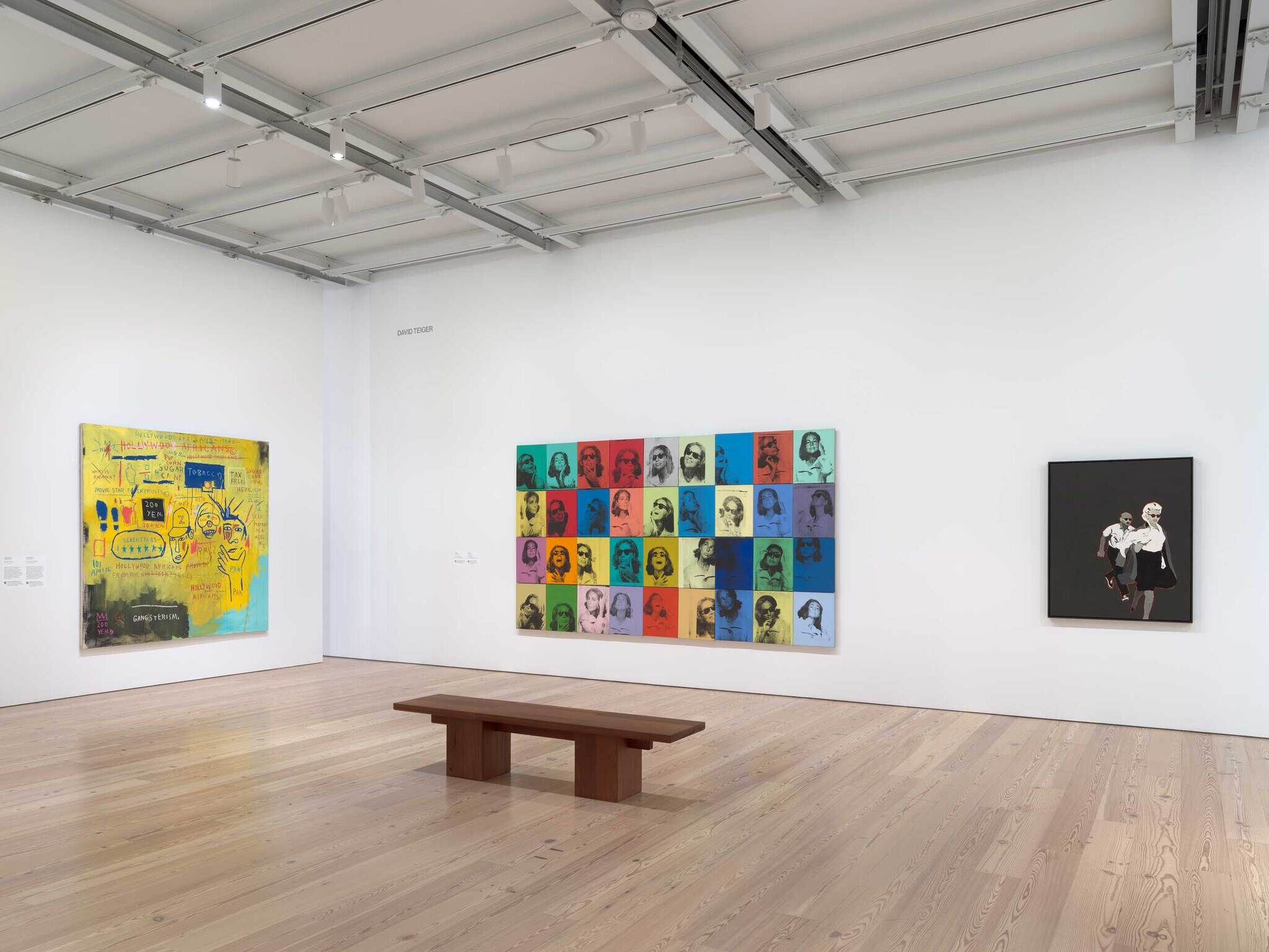 Art gallery room with three colorful modern paintings on white walls and a wooden bench in the center.