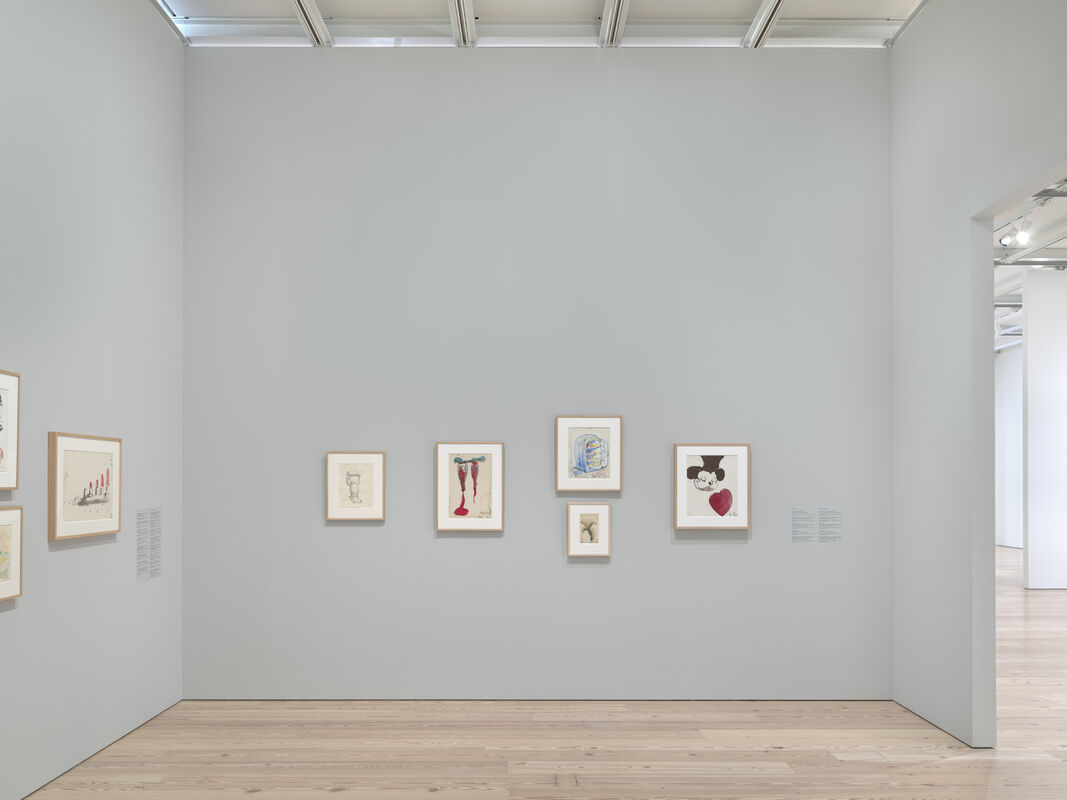 Several framed drawings and paintings hang on a light gray gallery wall, each featuring abstract or cartoon-like figures.