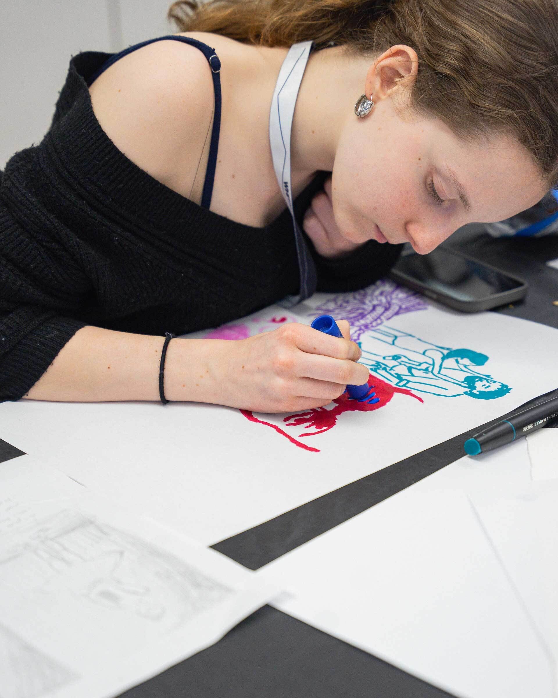 A young woman uses colored markers to draw people on white paper while leaning close to her artwork.