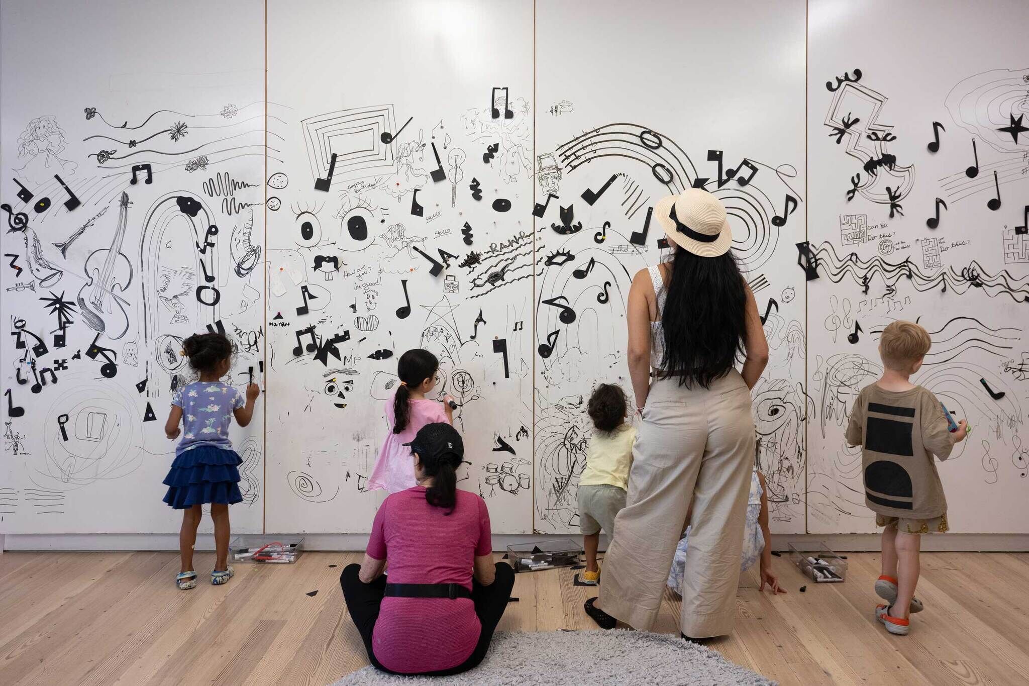 Children and adults draw and add musical notes to a large white wall covered in creative black marker art.