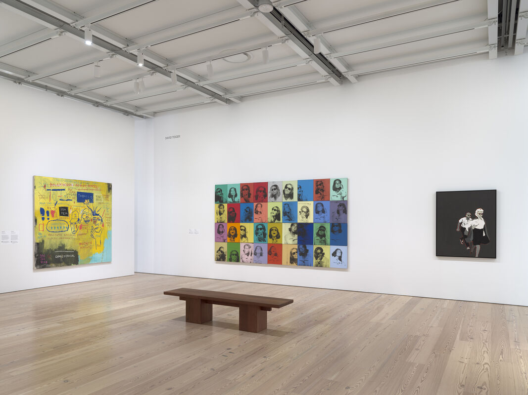 Three colorful modern art paintings hang on white walls in a gallery with a wooden bench in the center.