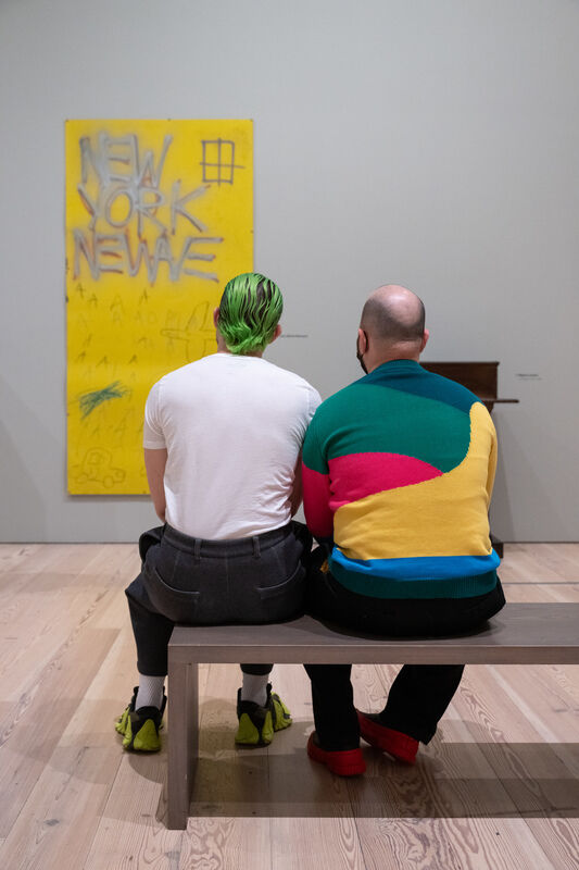 Two people sit on a bench facing a yellow artwork with the words "NEW YORK NEWAVE" written on it.