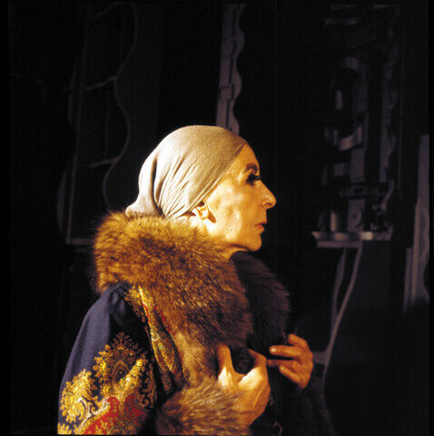 An older person wearing a headscarf and fur-collared coat stands in profile, hands holding the coat closed.