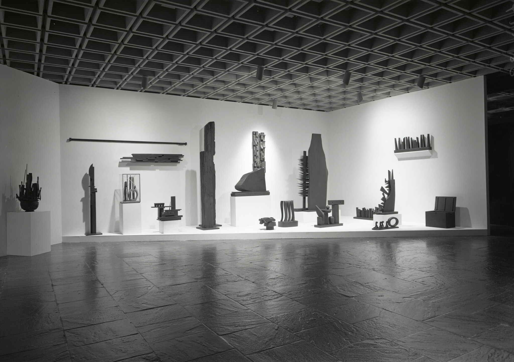 Several abstract metal and wood sculptures are displayed on white platforms against a white wall in a gallery.