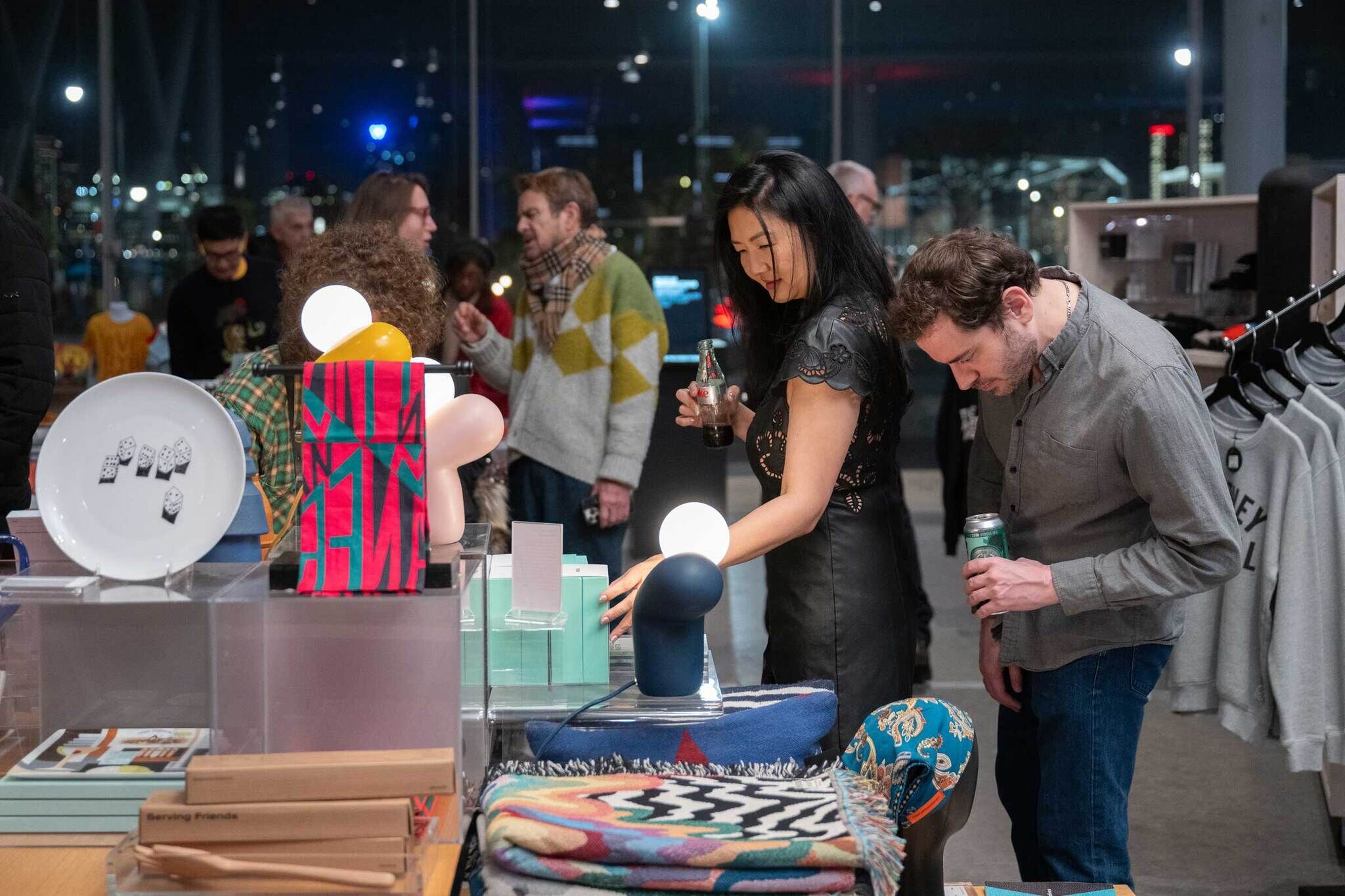 Two people look at items on a display table in a store, each holding a drink, with others nearby.
