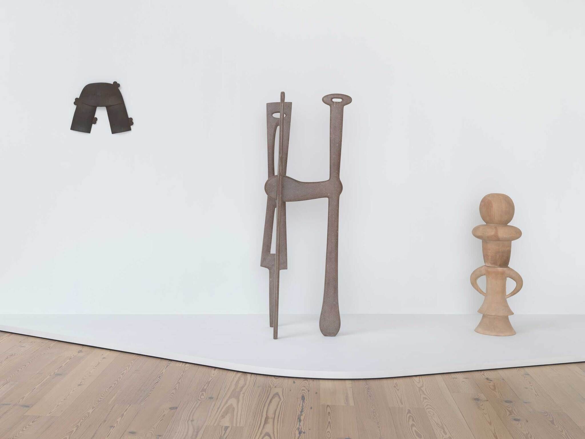 Three abstract sculptures stand against a white wall; one is mounted, two are on the floor with simple shapes.