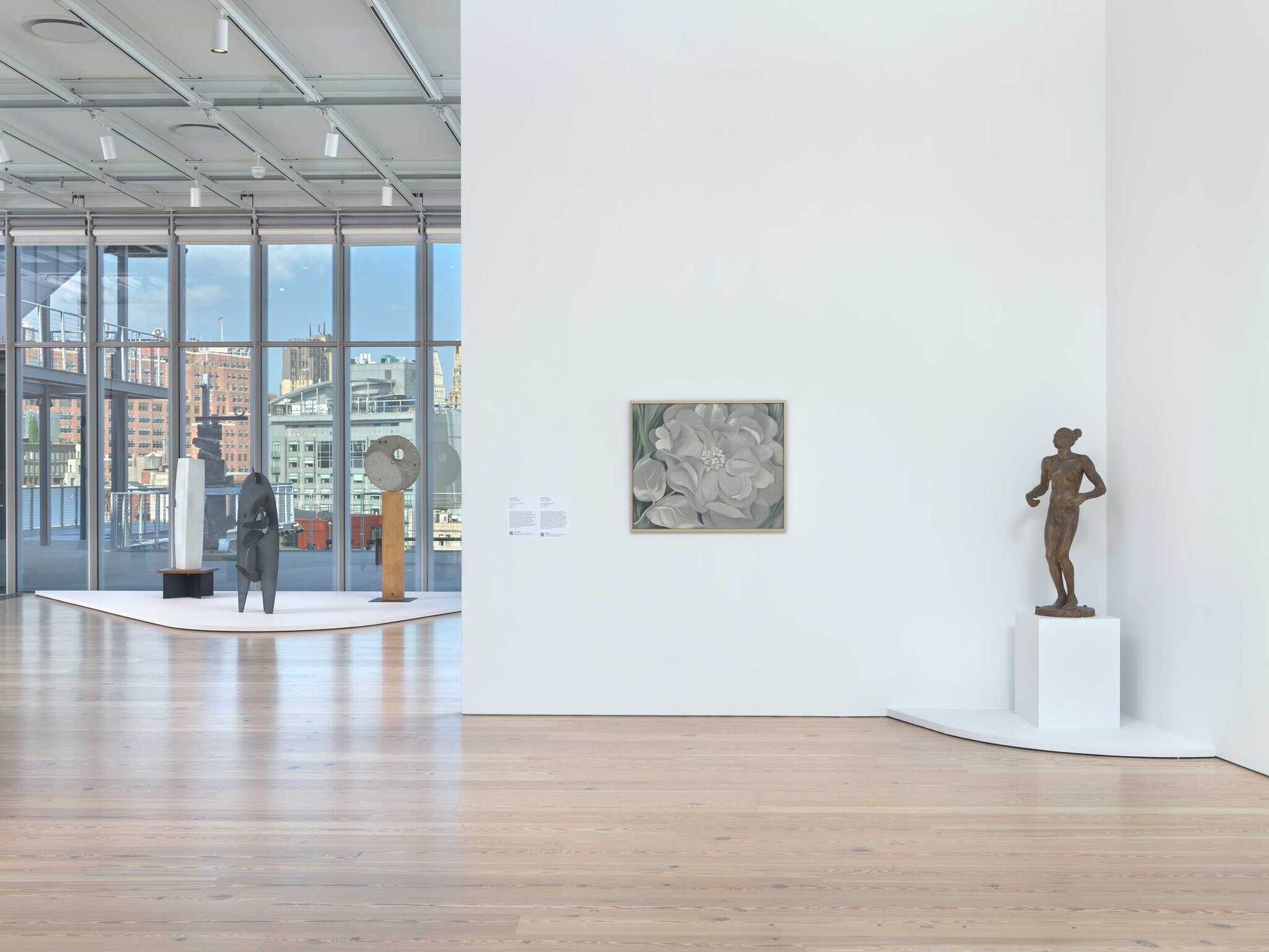 Art gallery with sculptures on pedestals and a painting of white flowers on a white wall. City buildings visible outside.