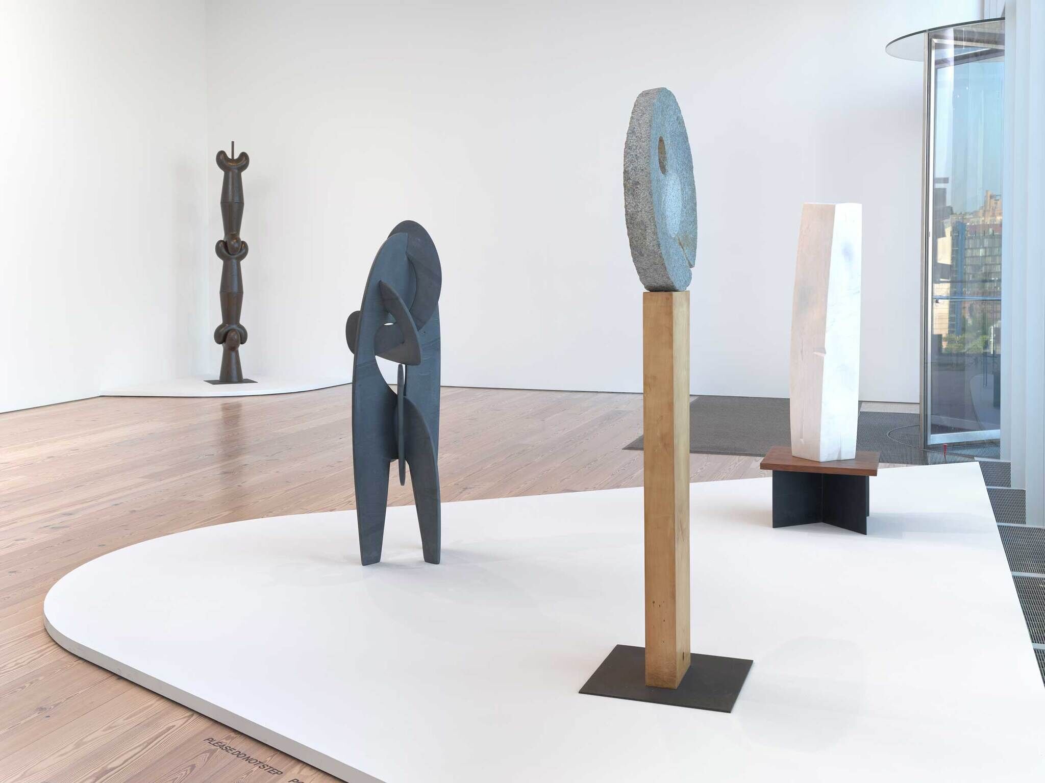 Four abstract sculptures made of metal, stone, and wood are displayed on white platforms in a modern gallery.