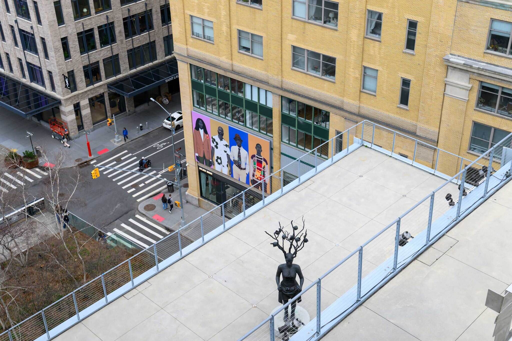 A rooftop with a statue of a person with tree branches for hair overlooks a city street and crosswalk.