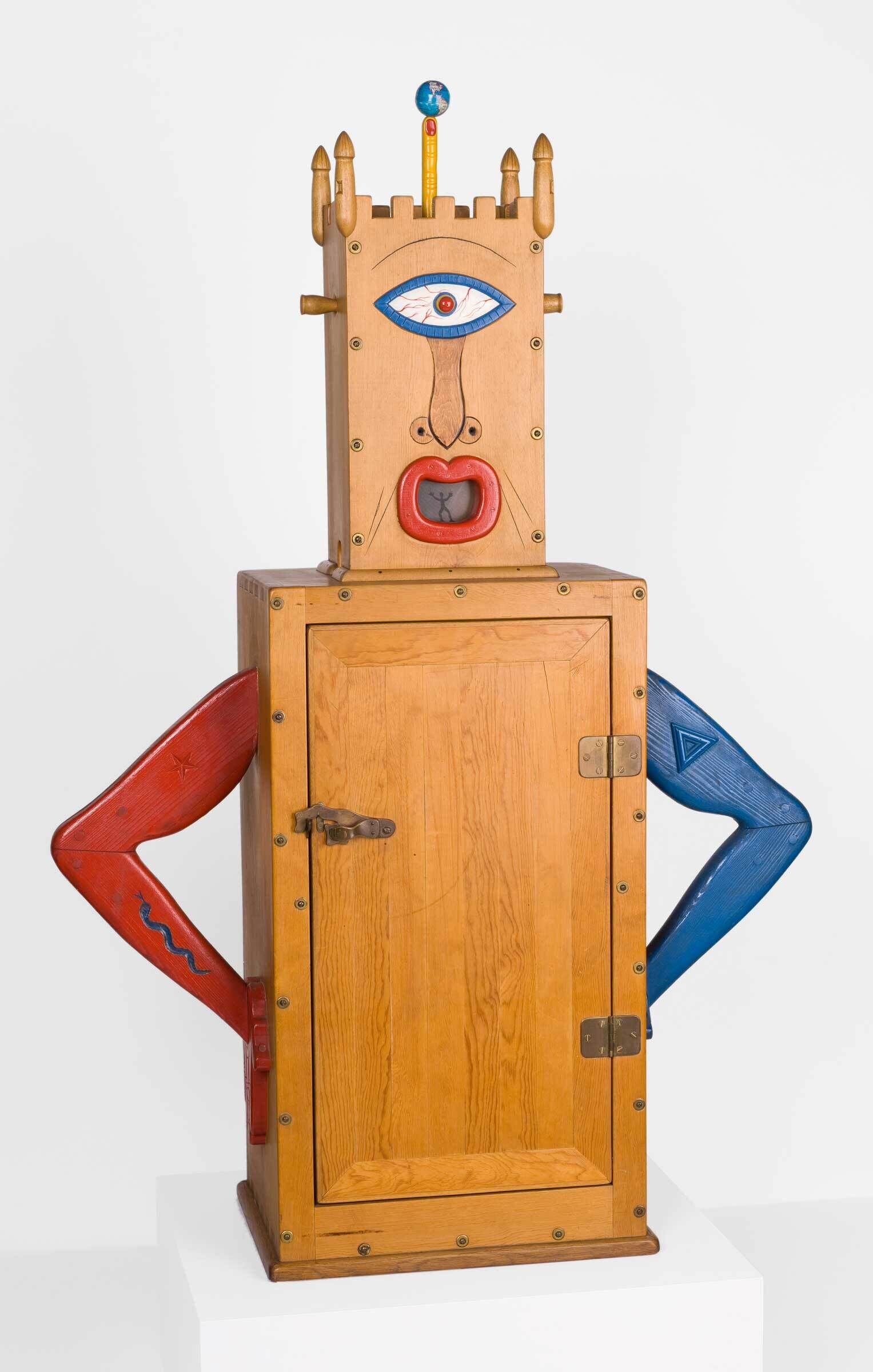 A wooden cabinet-like sculpture shaped like a stylized figure with red and blue painted arms bent at the hips, a rectangular body with a latched door, and a tall head resembling a castle tower. The head features a large carved eye, a protruding red mouth, and a small globe-topped rod extending upward. Made of pine with mixed media elements including metal, glass, and enamel. 