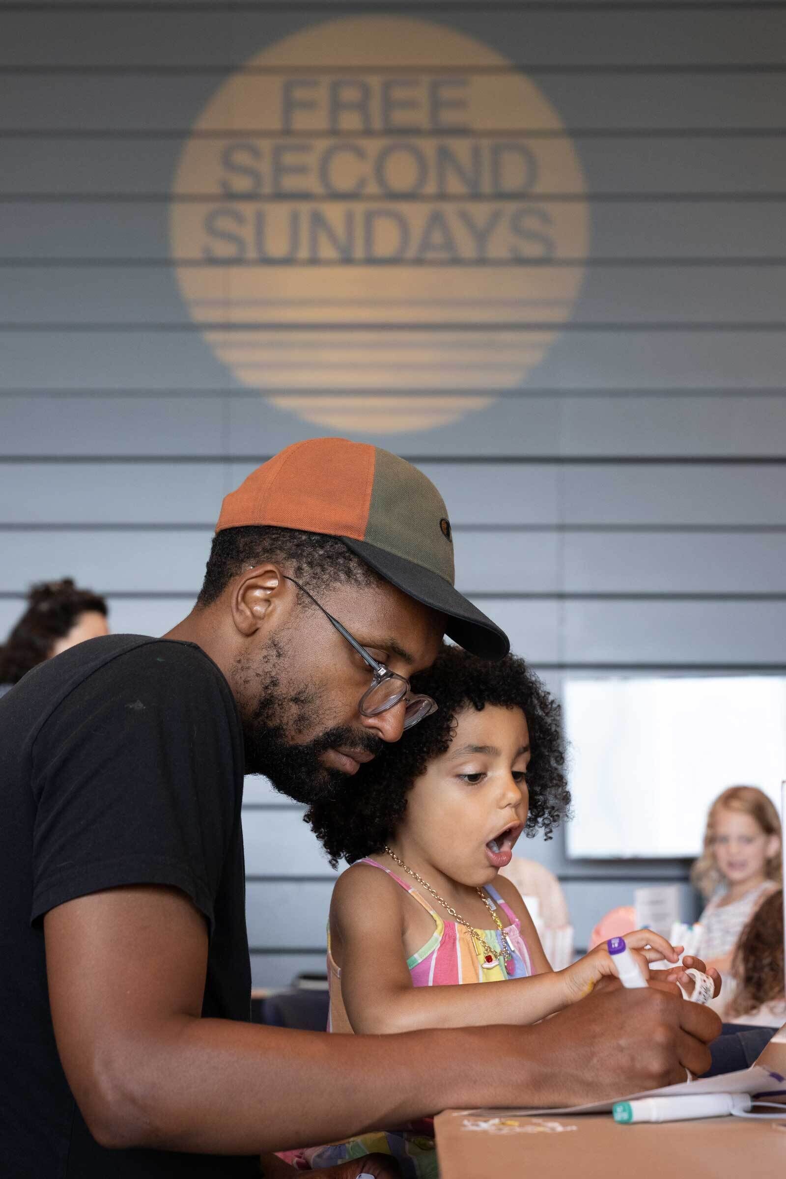 One adult helps a child with curly hair who is holding a marker, their mouth open in excitement or surprise. Behind them, other participants are also engaged in artmaking. On the wall, a large circular projection reads “FREE SECOND SUNDAYS.”  