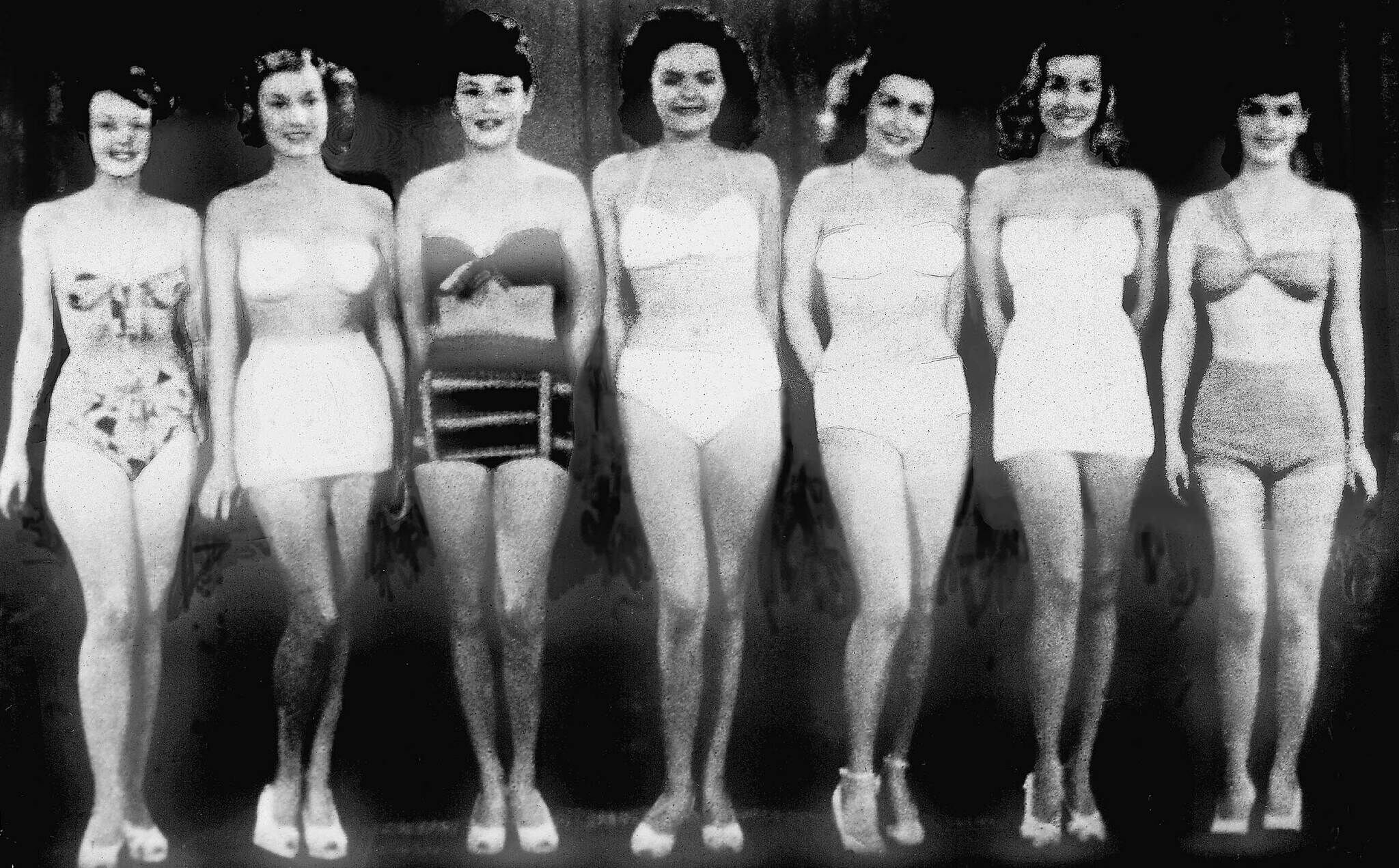 Seven women stand in a row wearing vintage swimsuits, smiling and facing forward against a plain background.