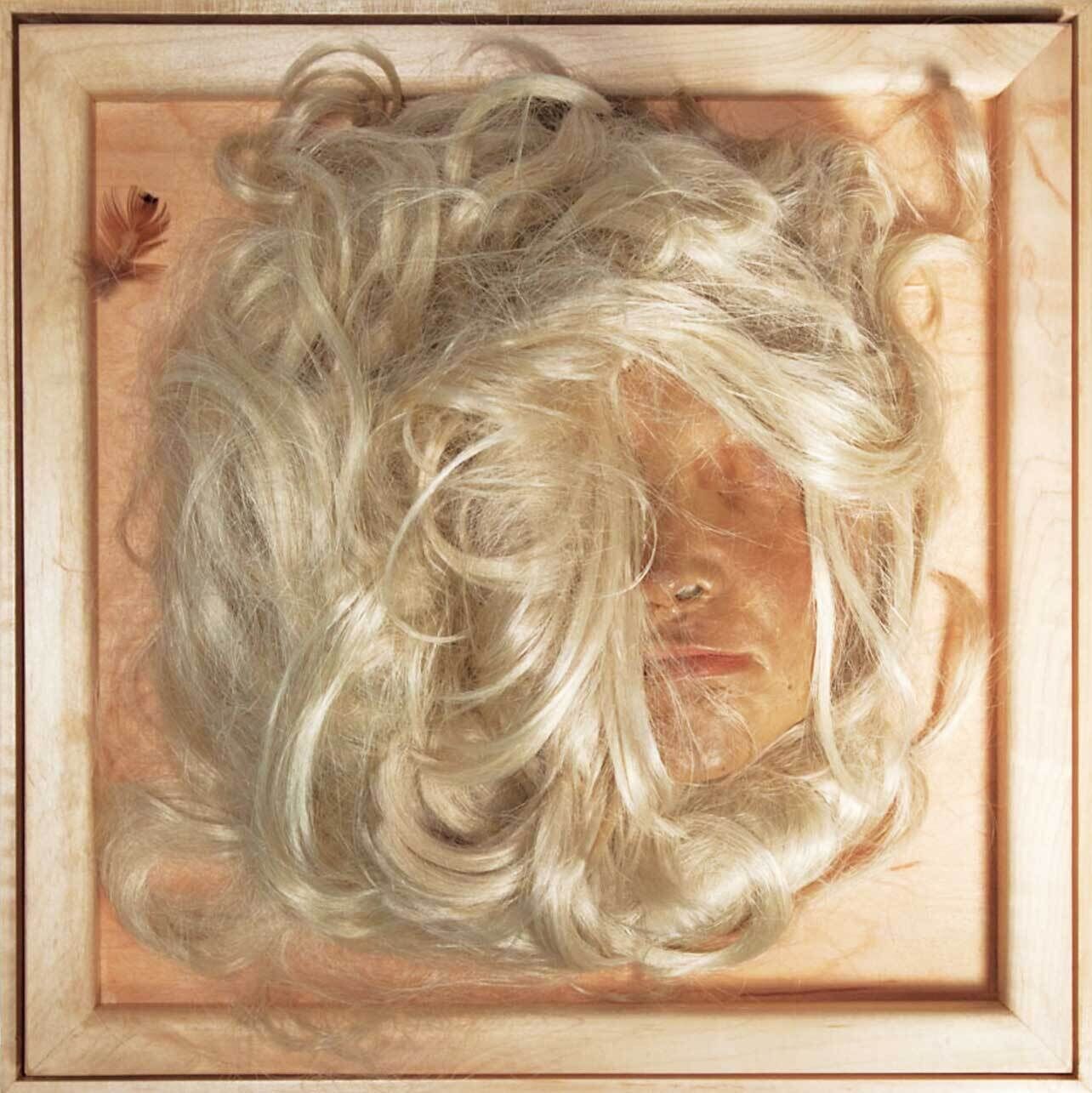 A framed display with a blonde wig covering a realistic face mask, and a small brown feather in the corner.
