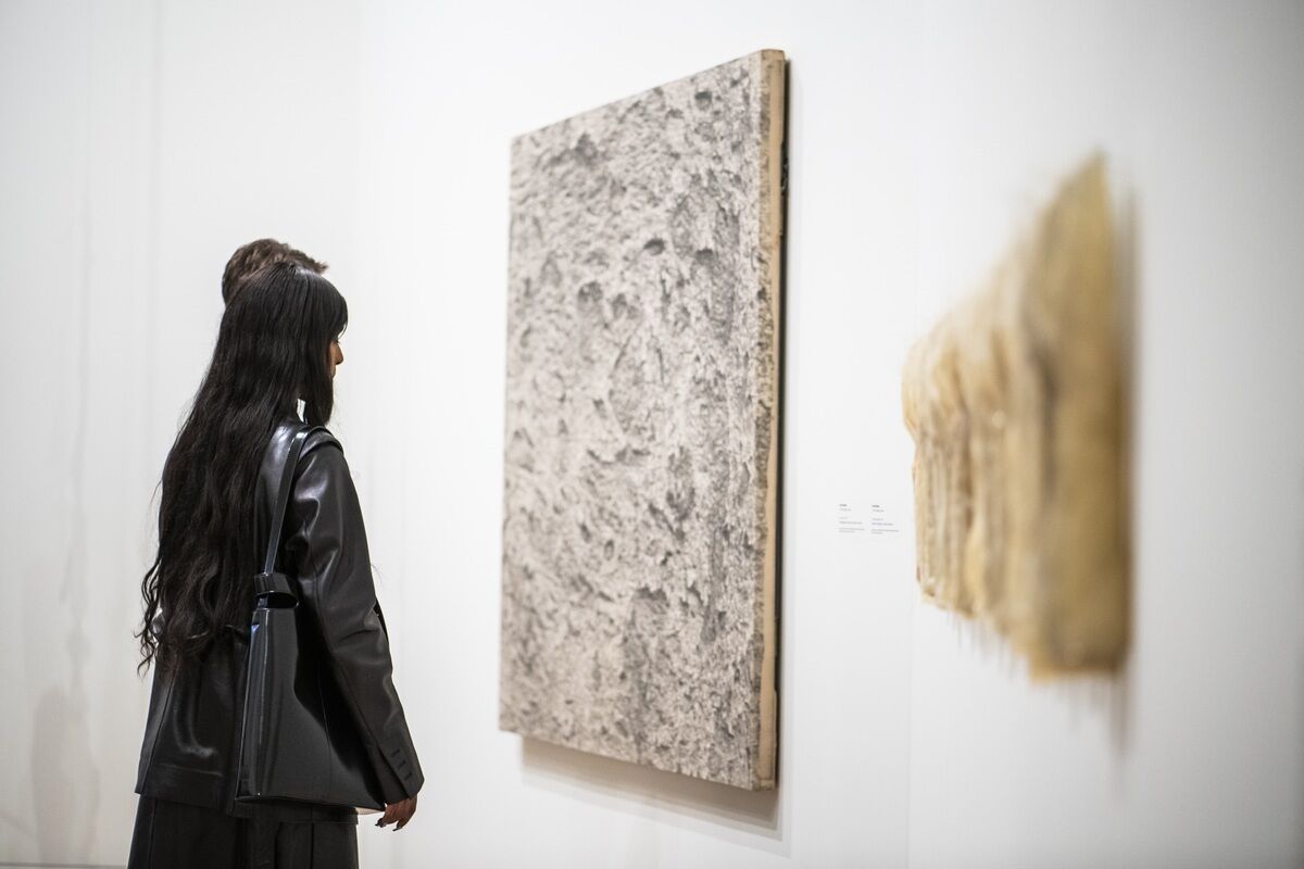 Two people observe textured artworks on a white gallery wall, one resembling a gray, rough surface and the other a soft, beige material with vertical elements.