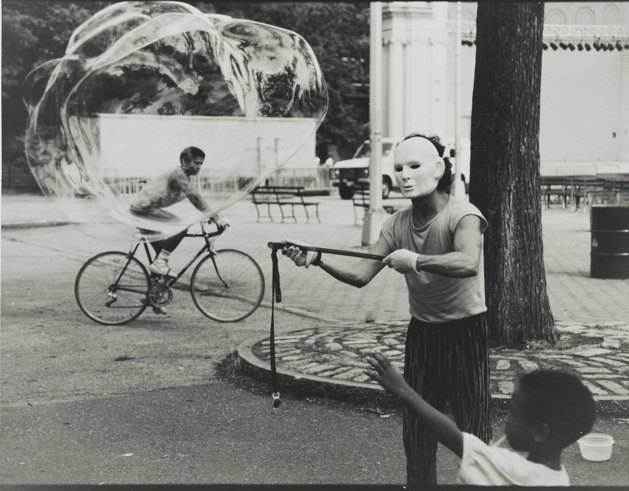 Person wearing a white mask creates a giant bubble while a child reaches out and a cyclist passes by.