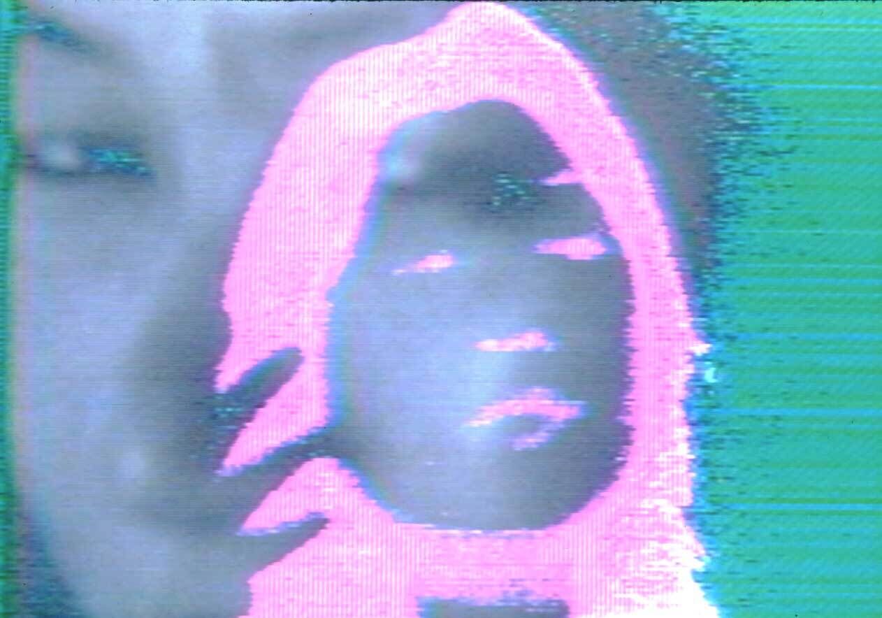 A person’s face appears on a screen with a pink digital outline and green background, creating a glitch effect.