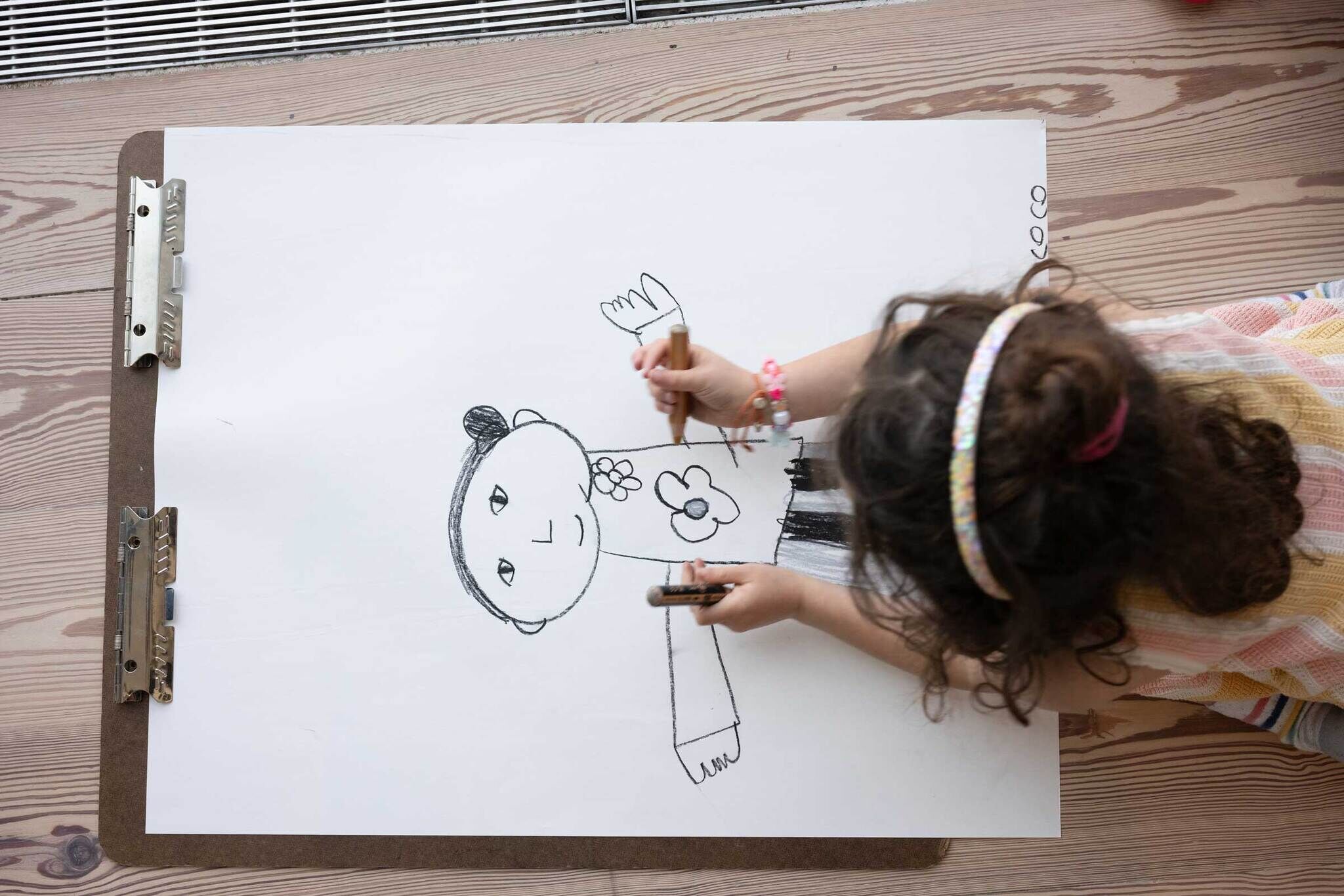 Young child with curly hair draws a person with a flower on their shirt using black crayons.