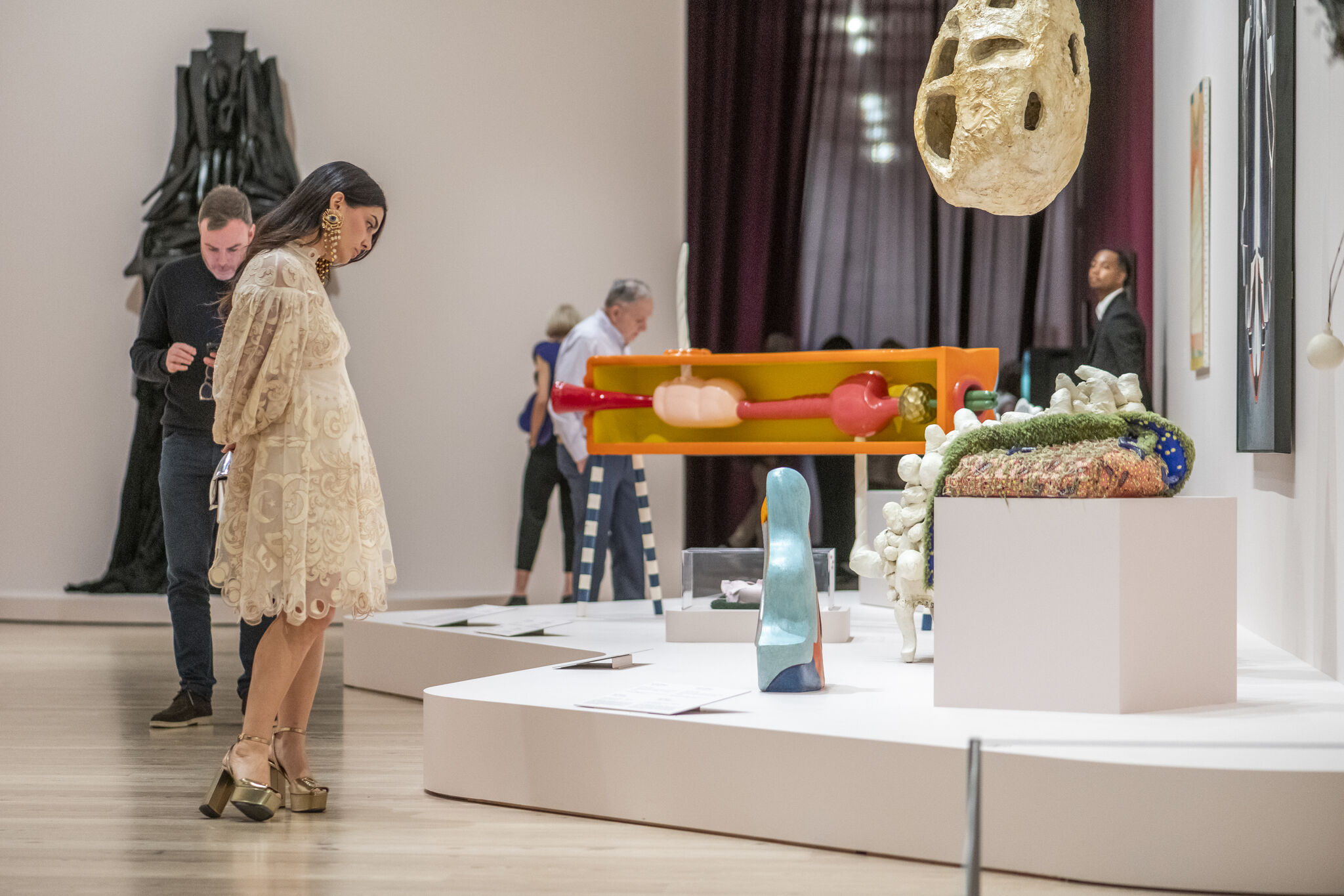 A museum visitor in a lace dress leans forward to closely examine colorful sculptures in a gallery.