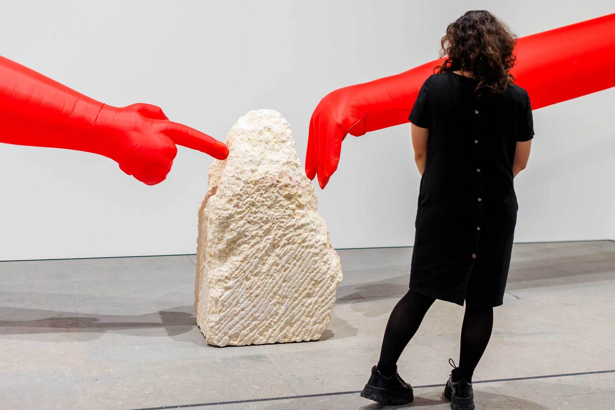 Person in black dress stands near a rough stone, with two large red inflatable hands pointing at it.