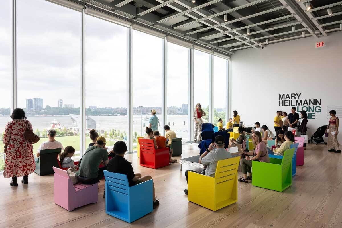 People sit on colorful chairs in a bright room with large windows; wall text reads "Mary Heilmann: Long Line."