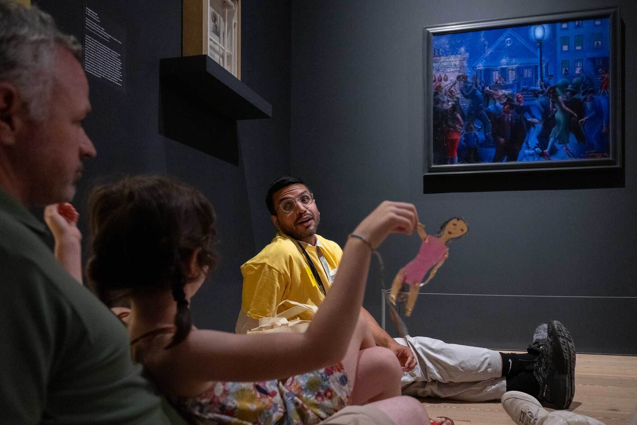 A man in a yellow shirt talks to a child holding a paper doll, sitting near a painting in a museum.